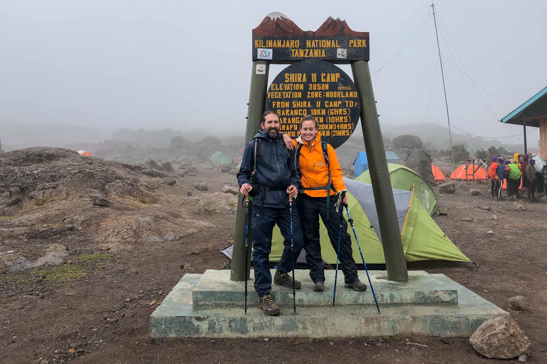 A man in blue and a woman in orange standing at the brown "Shira II Camp" Kilimanjaro sign with tents and people in the background shrouded in mist 
