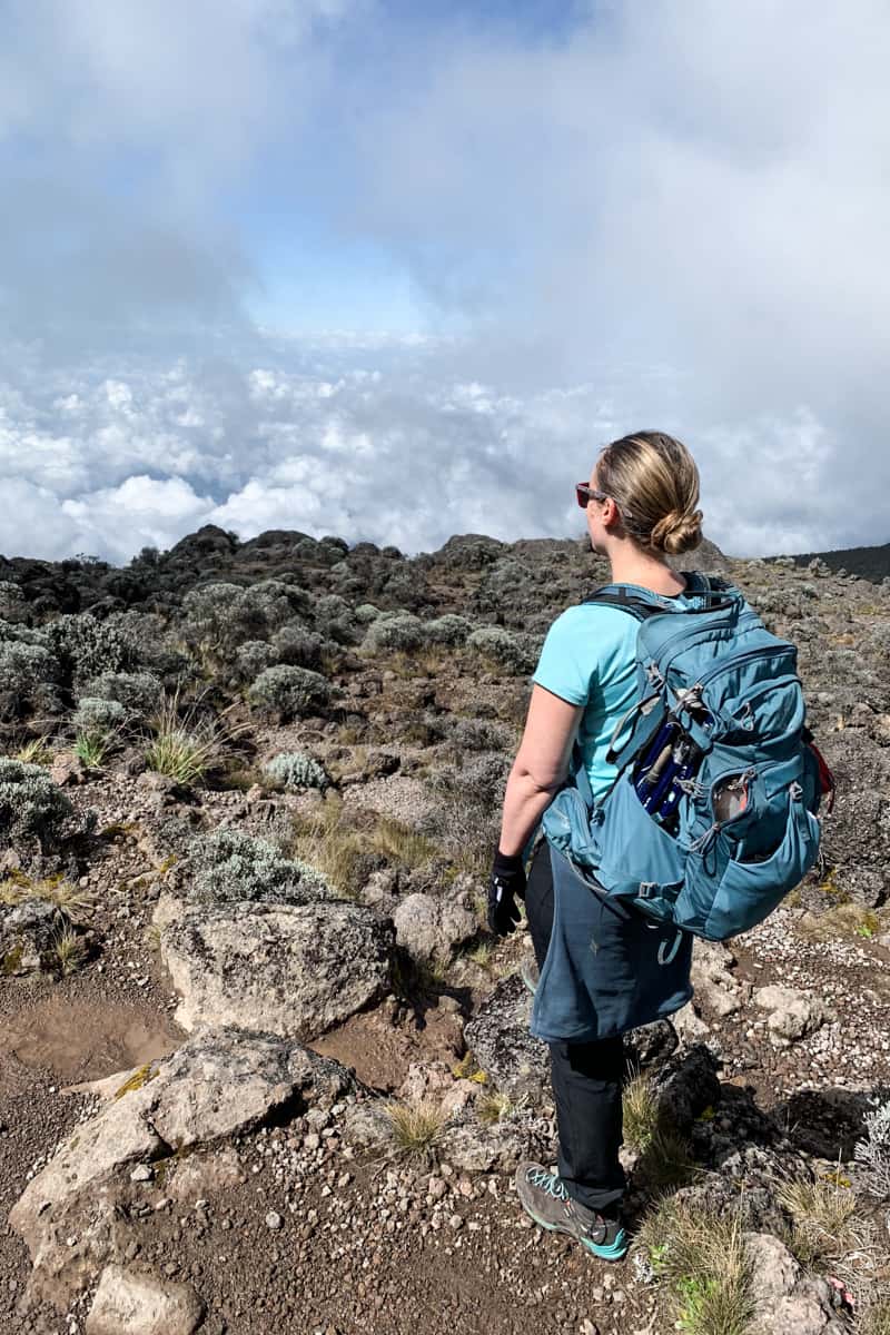 A woman in a blue t-shirt, with a blue daypack stands on a rocky path overlooking a Kilimanjaro valley covered in clouds
