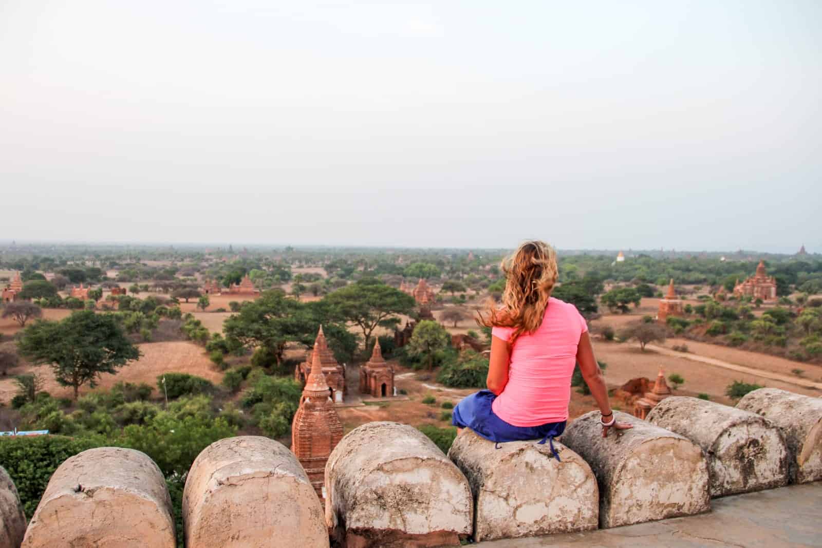 A woman in a neon pink t-shirt and purple pants sits on an arched stone watching sunset from the top of a temple in Bagan, Myanmar. In the fields ahead are a scattering of golden brown temples poking through the grass. 