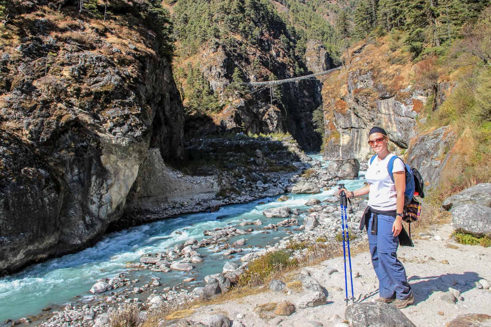 A woman in t-shirt and trekking pants holds a walking pole while standing next to a bright turquoise river in a small rocky canyon in the Nepalese Himalayas