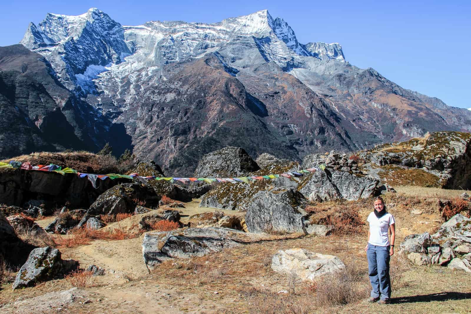 A woman in a white t-shirt and blue trekking pants stands in front of large black rocks strewn with multi-coloured prayer flags and a huge snow capped mountain face