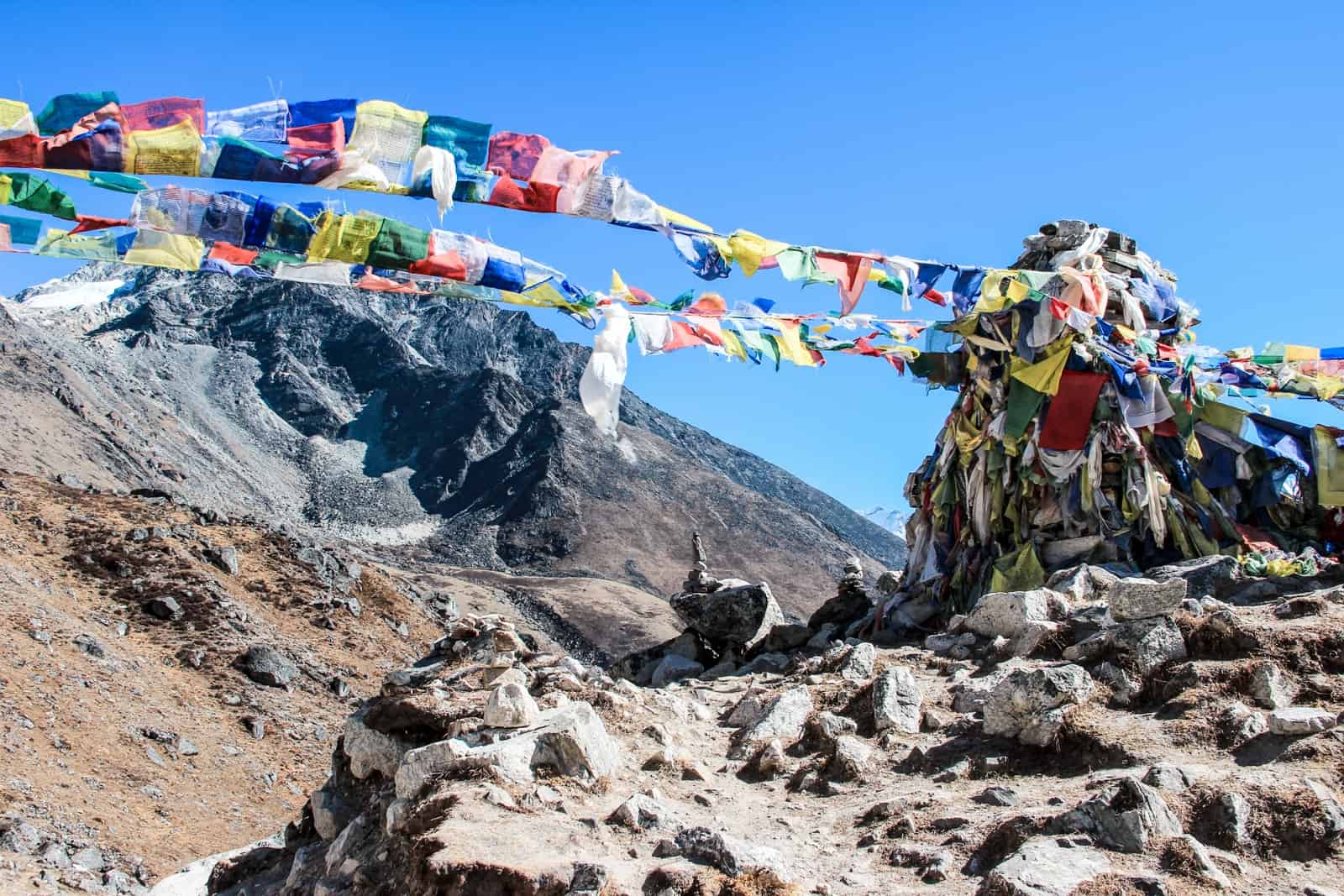 Two lines of multi-coloured prayer flags connect to a tall pile of rocks in a mountain valley on the Everest Base Camp trekking route