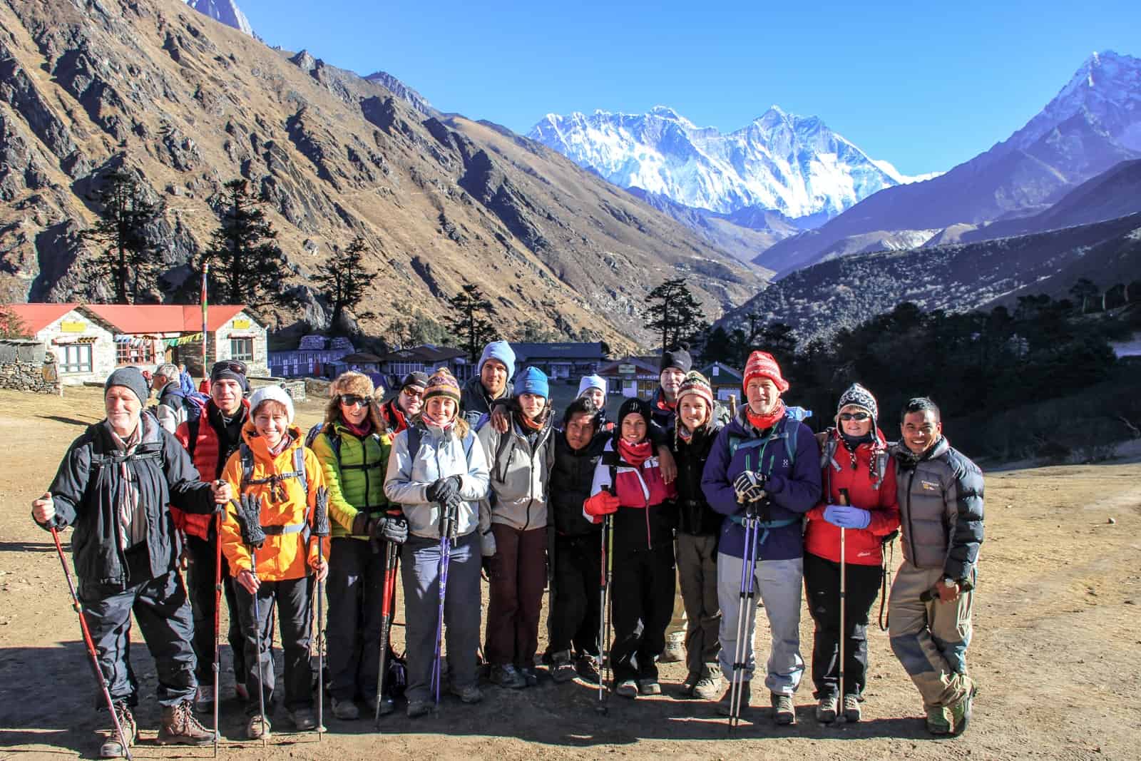 15 Everest Base Camp trekkers stand alongside their guide on the far right, outside a small complex of Nepali teahouses, backed by snow capped mountains