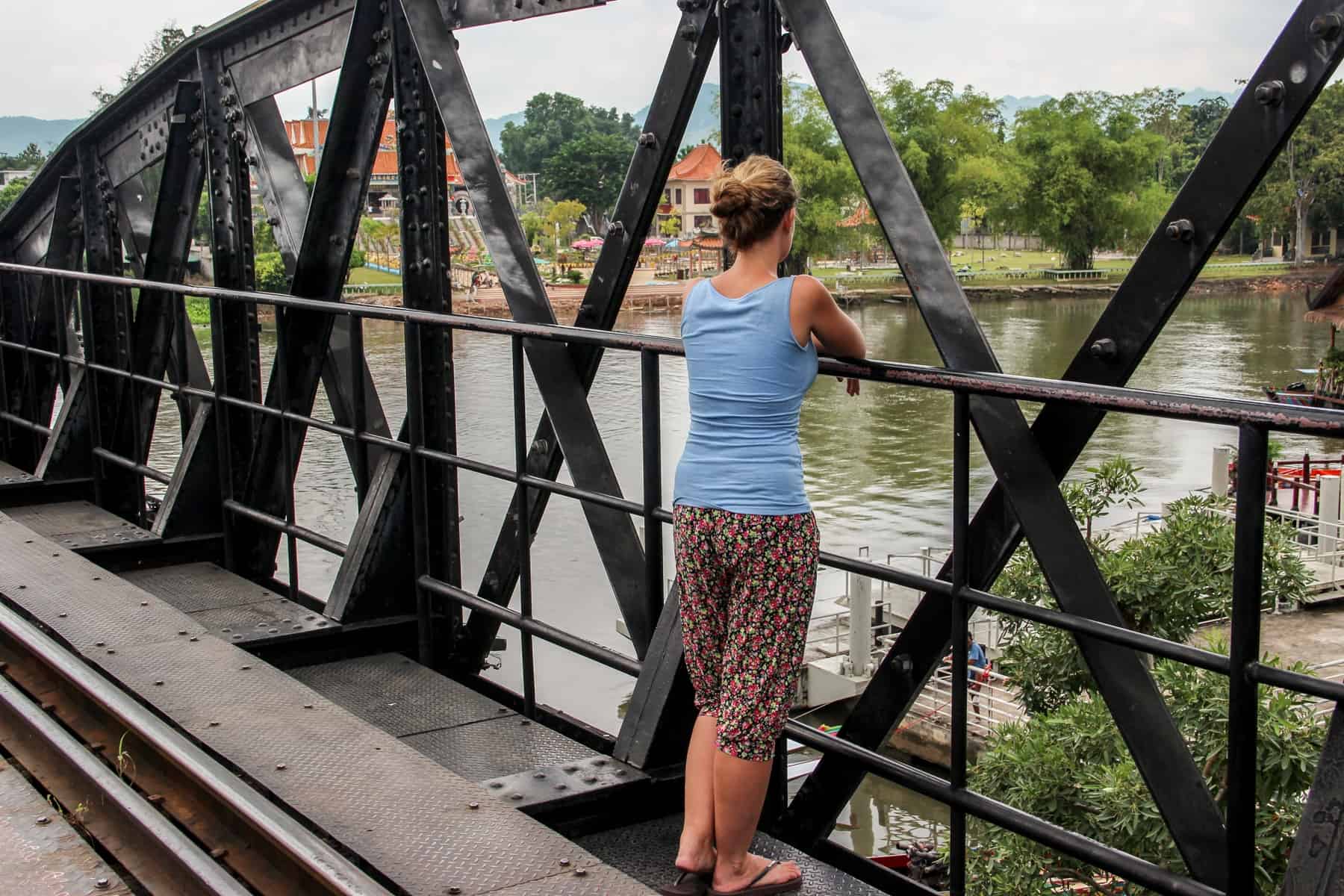 A woman leans on black rails on a platform on the Bridge over the River Kwai looking out towards the Thai river, greenery and orange roofed buildings. 