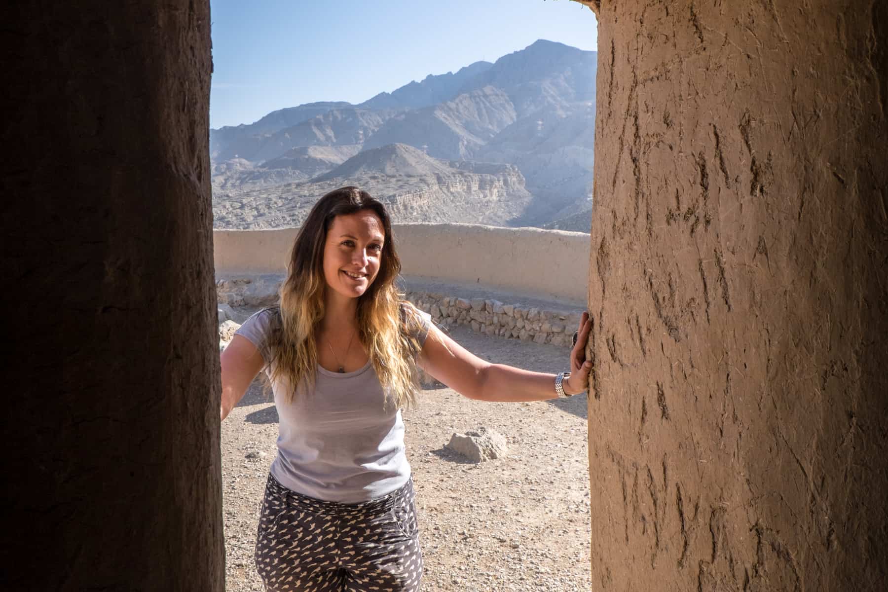 A woman stands in a doorway of an historic fort in Ras Al Khaimah made of mud clay. Behind her is a triangular mountain range
