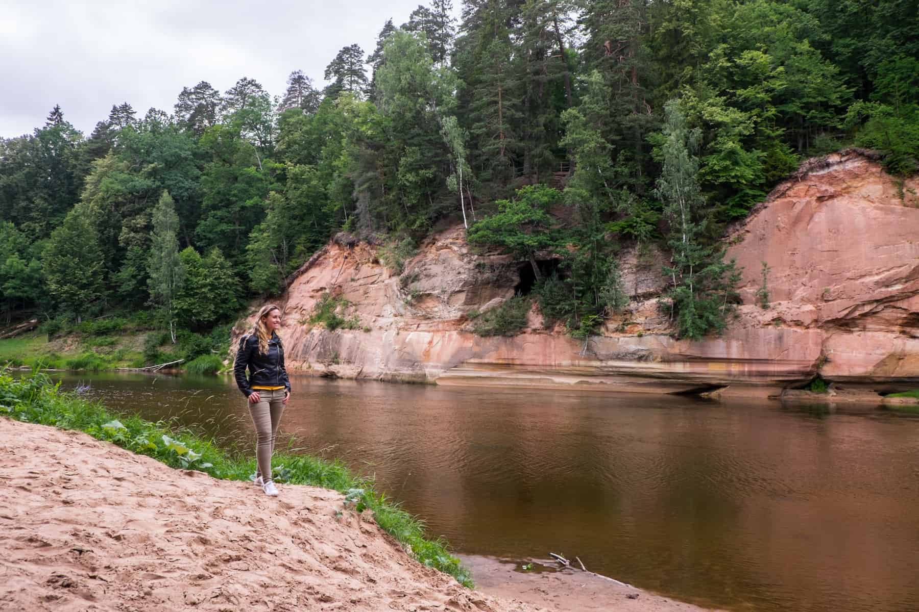 A woman in khaki pants and a black jacket stands on a sandy riverbank looking towards an ochre red cave under a green forest