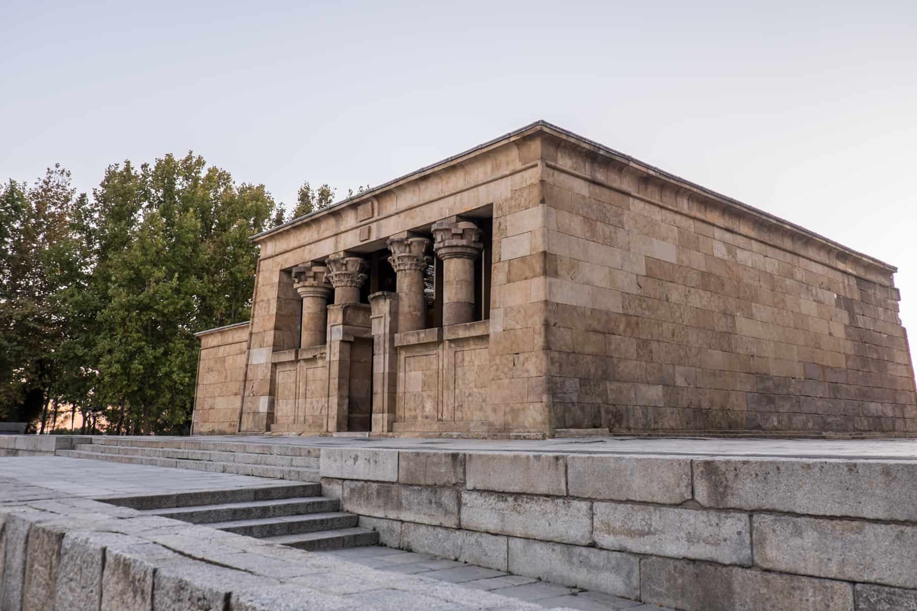 The golden stoned rectangular Egyptian Temple of Debod in Madrid, with four stone columns. The structure stands upon a surface of white stones, with a staircase. 