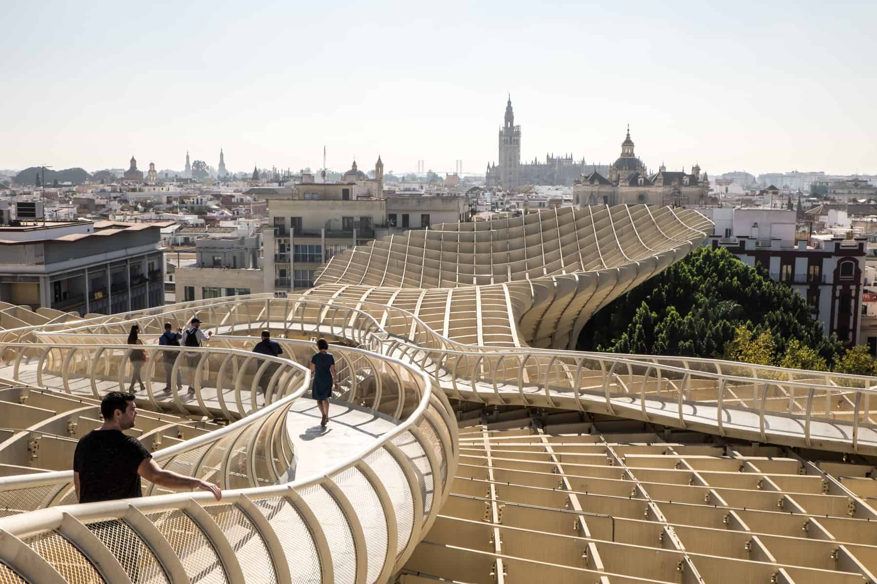 The cream slatted, criss-cross design of the modern artwork in Seville known as Metropol Parasol. These mushroom shaped structures, which people are walking through on curved platforms are elevated to sit in line with the skyline that spreads behind it. 