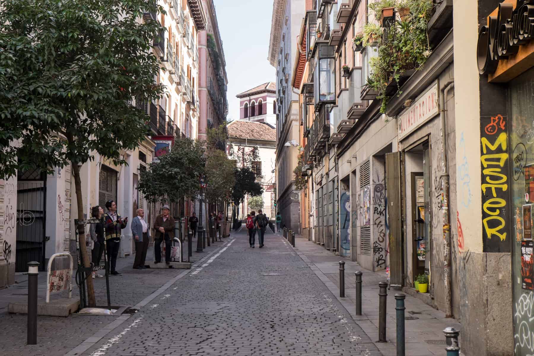 A street in Madrid whose mostly beige stone walls are covered in graffiti. Two people are walking down the street, while four people stand to one side talking. 