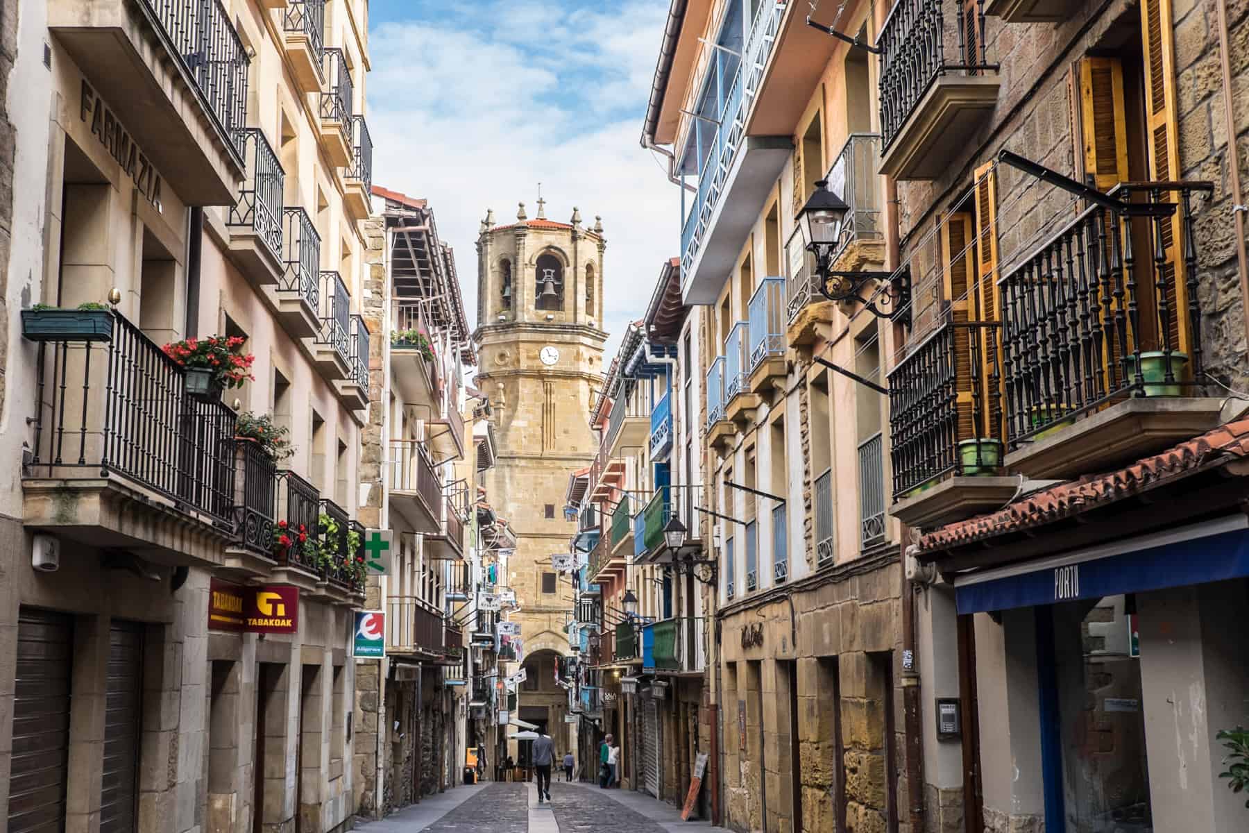A golden church tower stands at the end and in the middle of an old stone building street full of balconies in the Spanish city of San Sebastian. A man is walking towards the tower. 