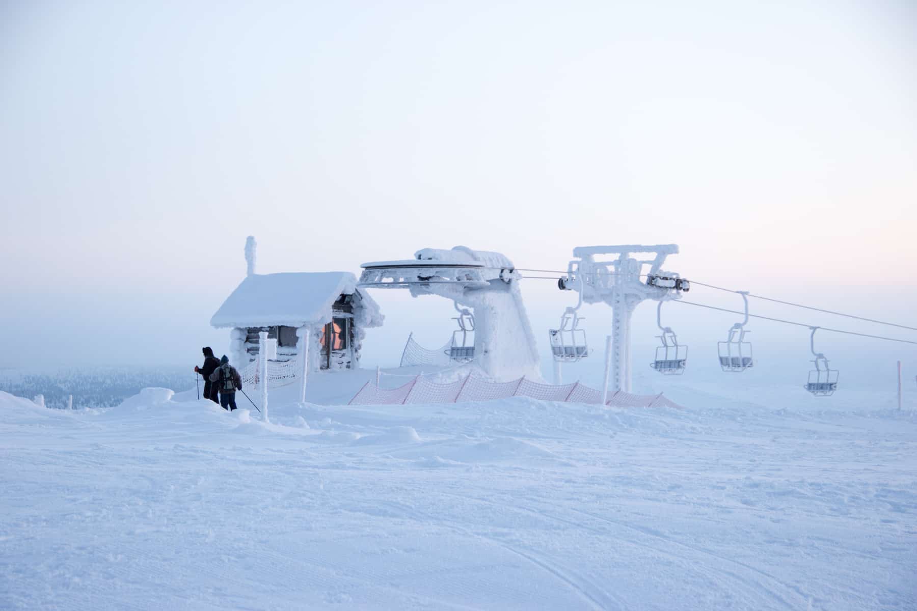 Two skiers dressed in black stand out against the scene of pure white at the top of a ski slope, next to a small ski lift, in Finnish Lapland. 