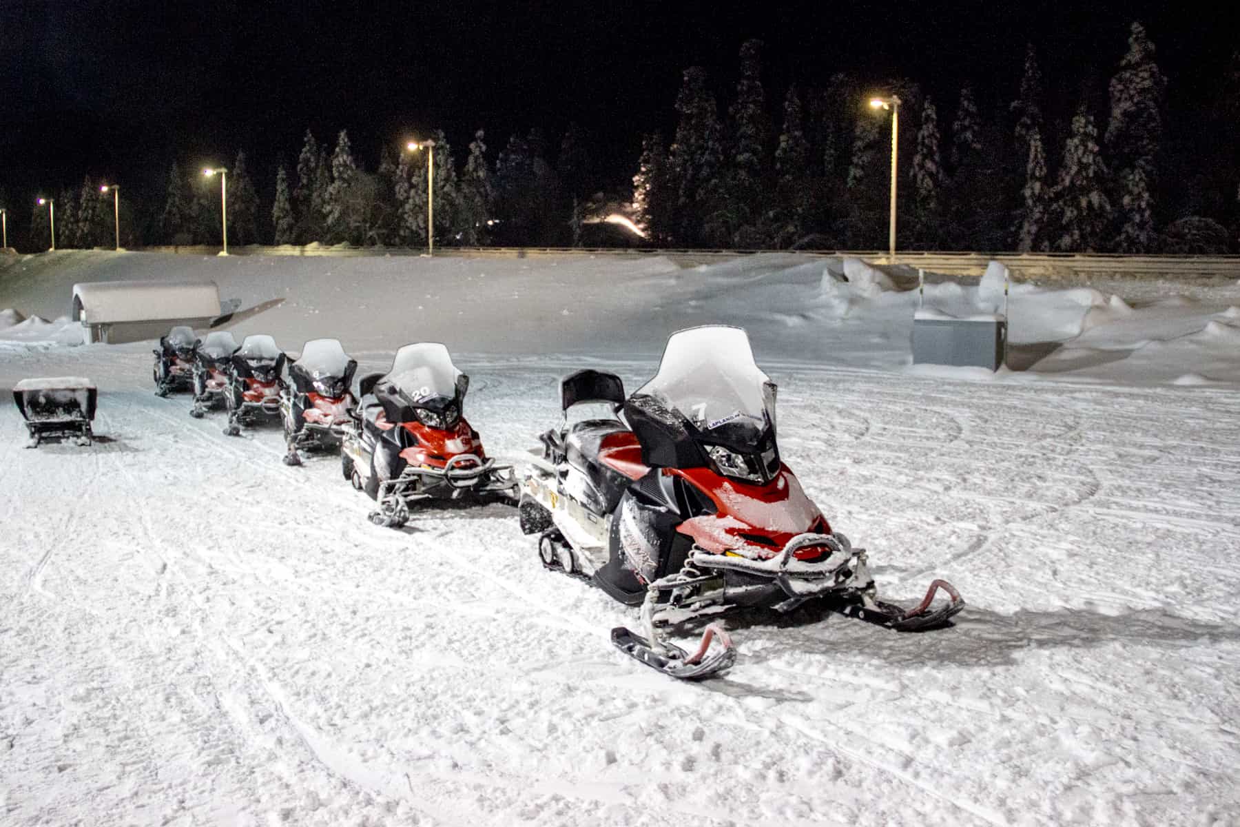 A row of six black and red snowmobiles on a snowy path under a black sky. 