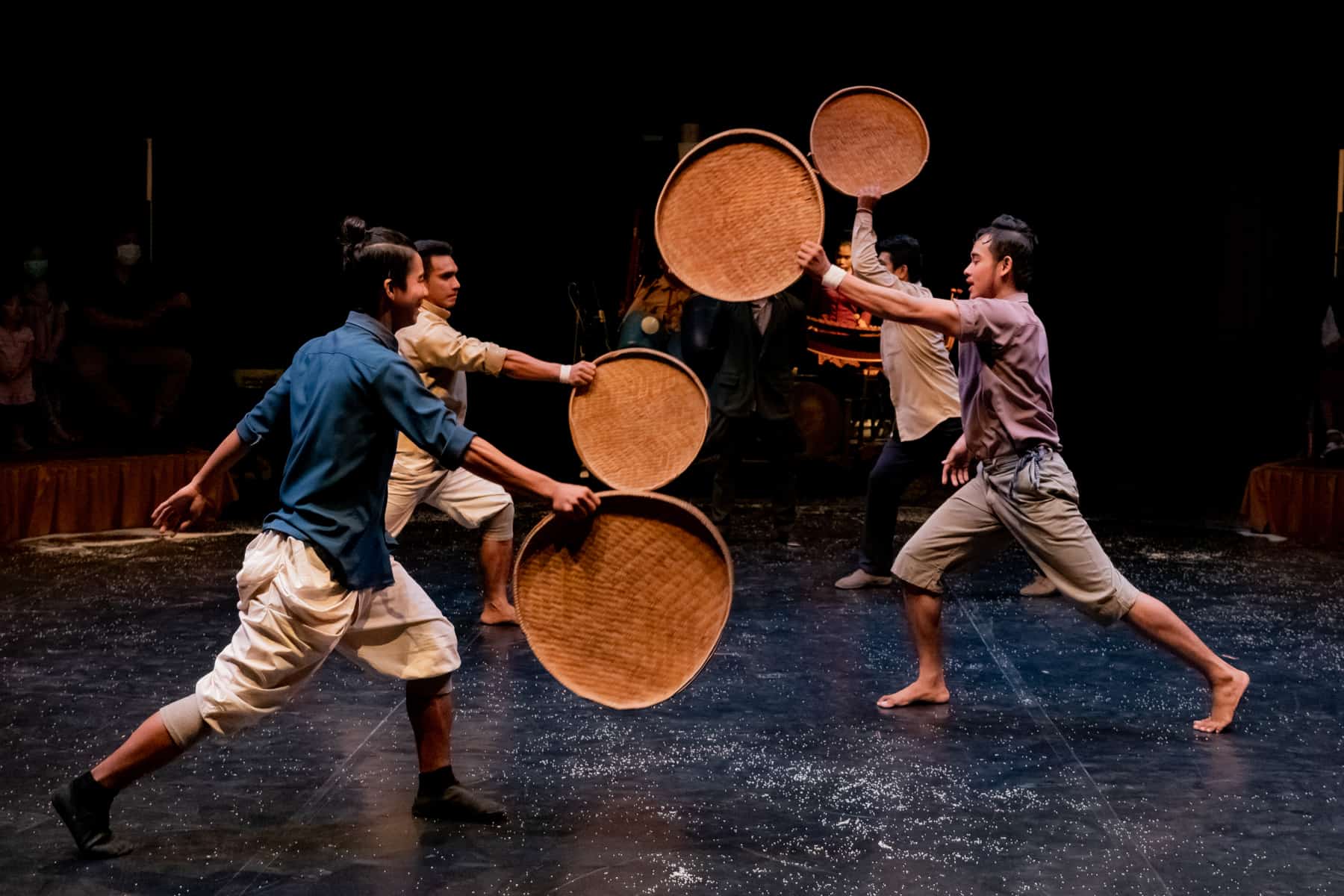 Four men perform a dance with woven rice trays in the Siem Reap Circus. The lighting in dark and musicians can be seen in the background. 