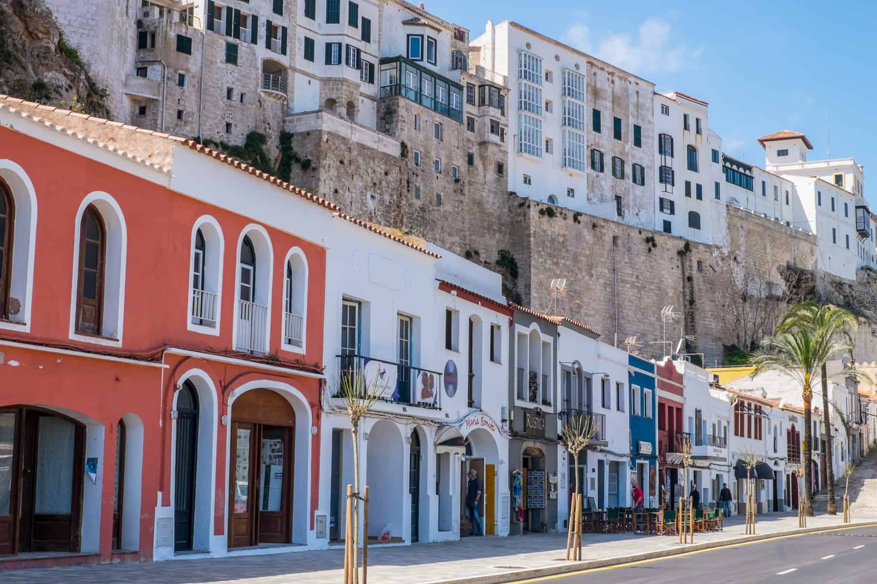 A street level, sloping row of houses painted white, blue and red in Port Mahon on the Balearic Island of Menorca.