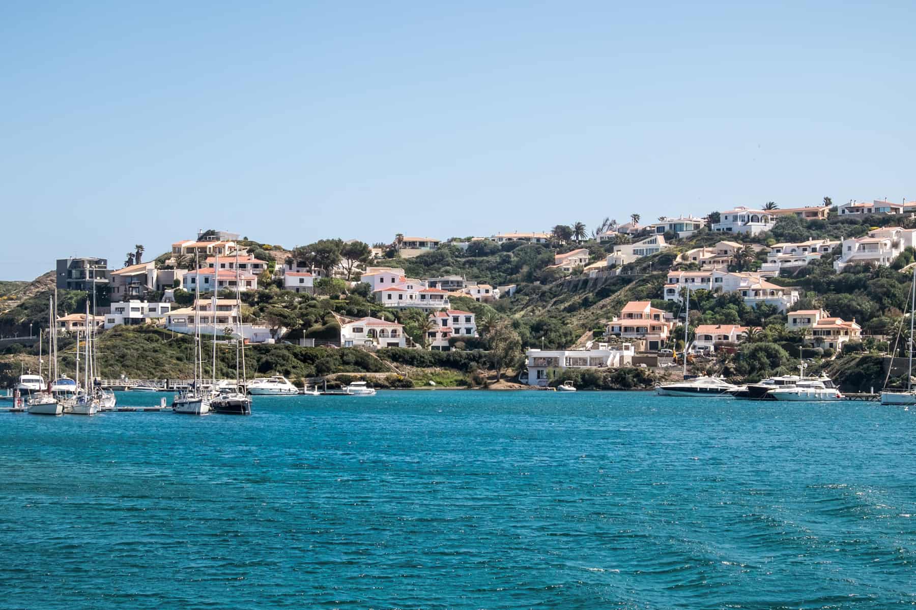 Small sailing boats on the bright blue waters of the Menorcan coastline, where rugged hilltops are covered in pretty white houses.