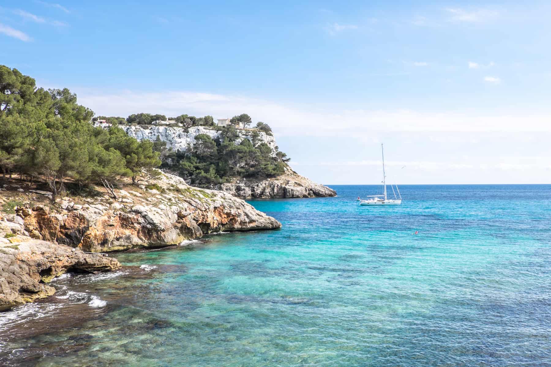 A single white sailing boat off the white cliff and rocky coastline of Menorca in Spain. 