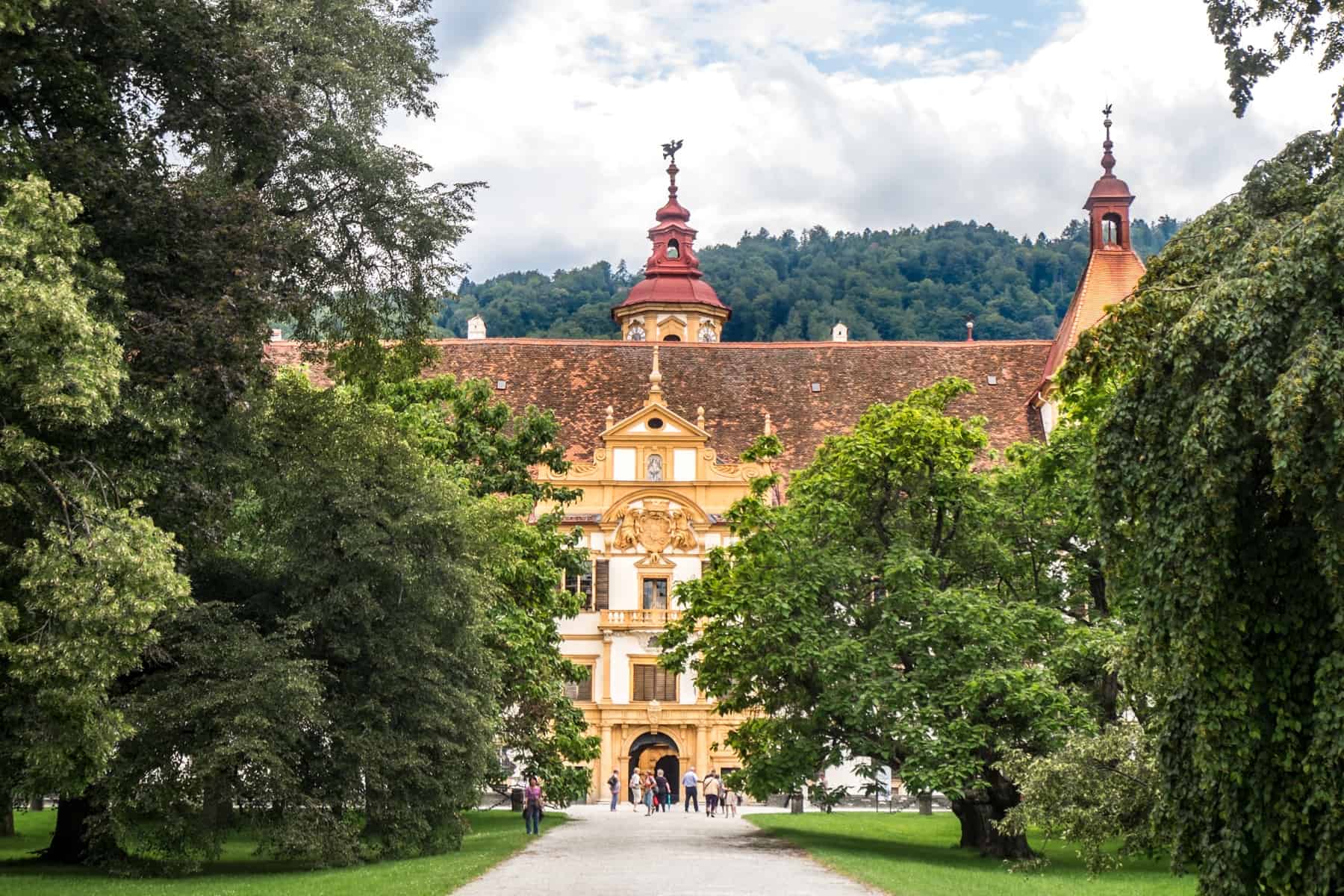 A pathway between think trees leads to an opulent golden yellow, red-roofed building - the entrance to the UNESCO World Heritage Site of Schloss Eggenberg in Graz