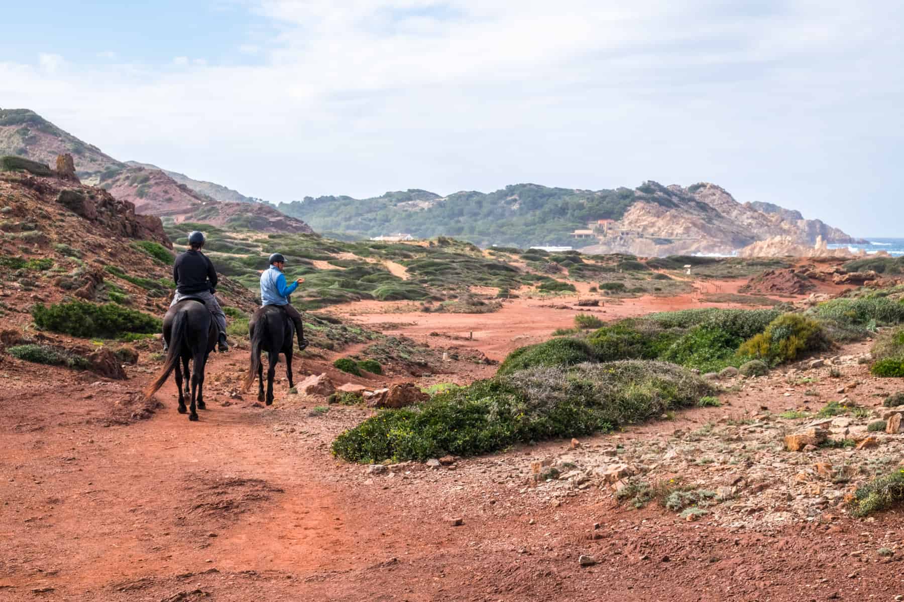 Two men on horseback trot through a rocky landscape with ochre red soil and patches of green foliage. 