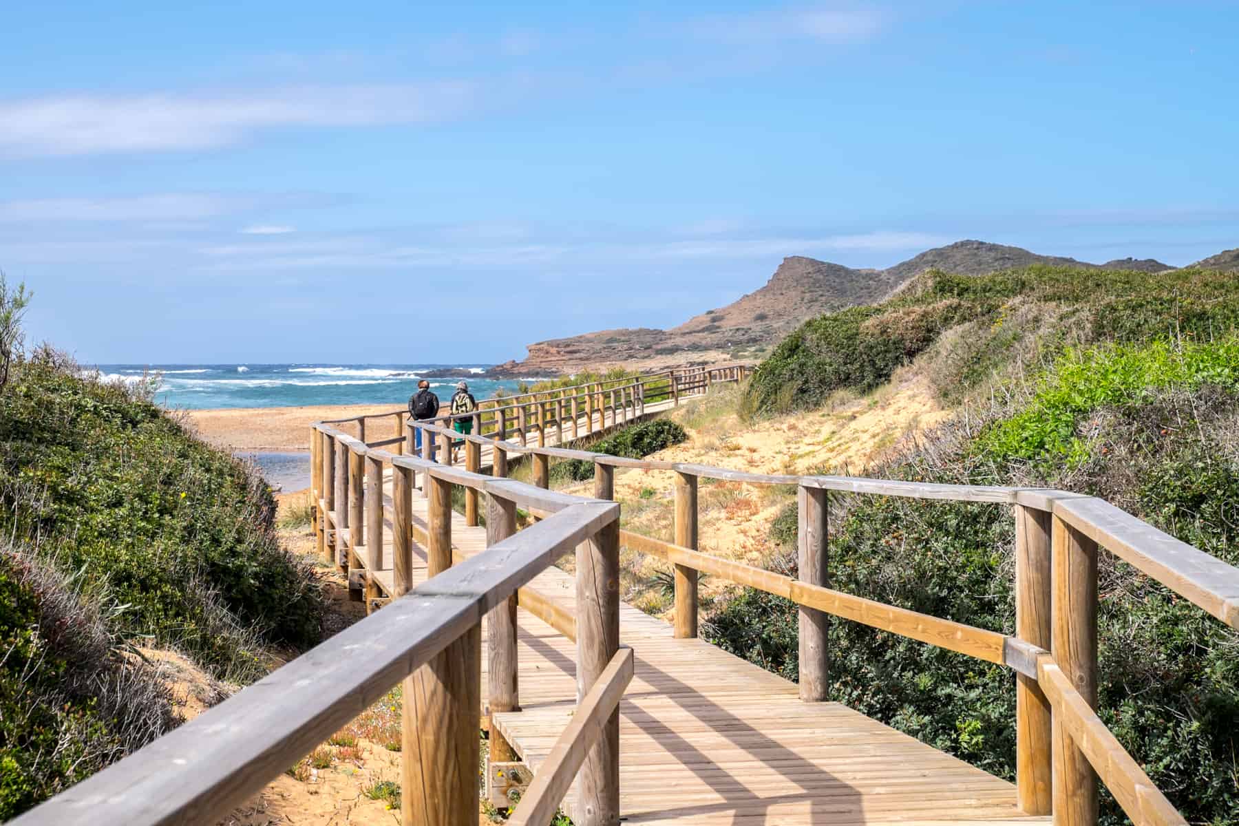 Two people walking the Cami de Cavalls in Menorca on an elevated wooden walkway over a yellow sand beach, backed by grassy hills. 