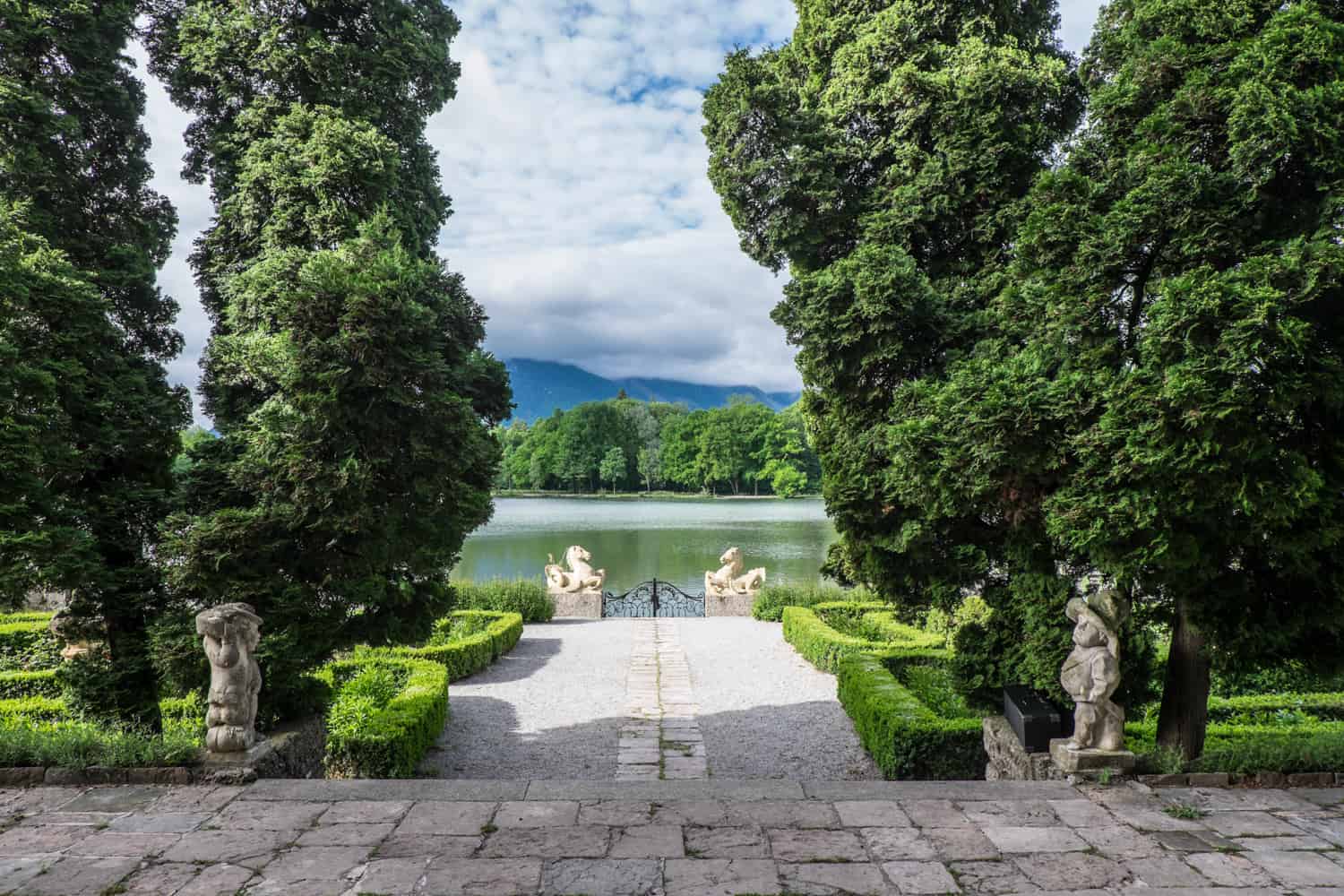 Two white stone horses' heads either side of a black metal gate overlooking a lake surrounded by trees. The 'Horse Head Gate' was made famous in The Sound of Music and is found at Schloss Leopoldskron in Salzburg, Austria.