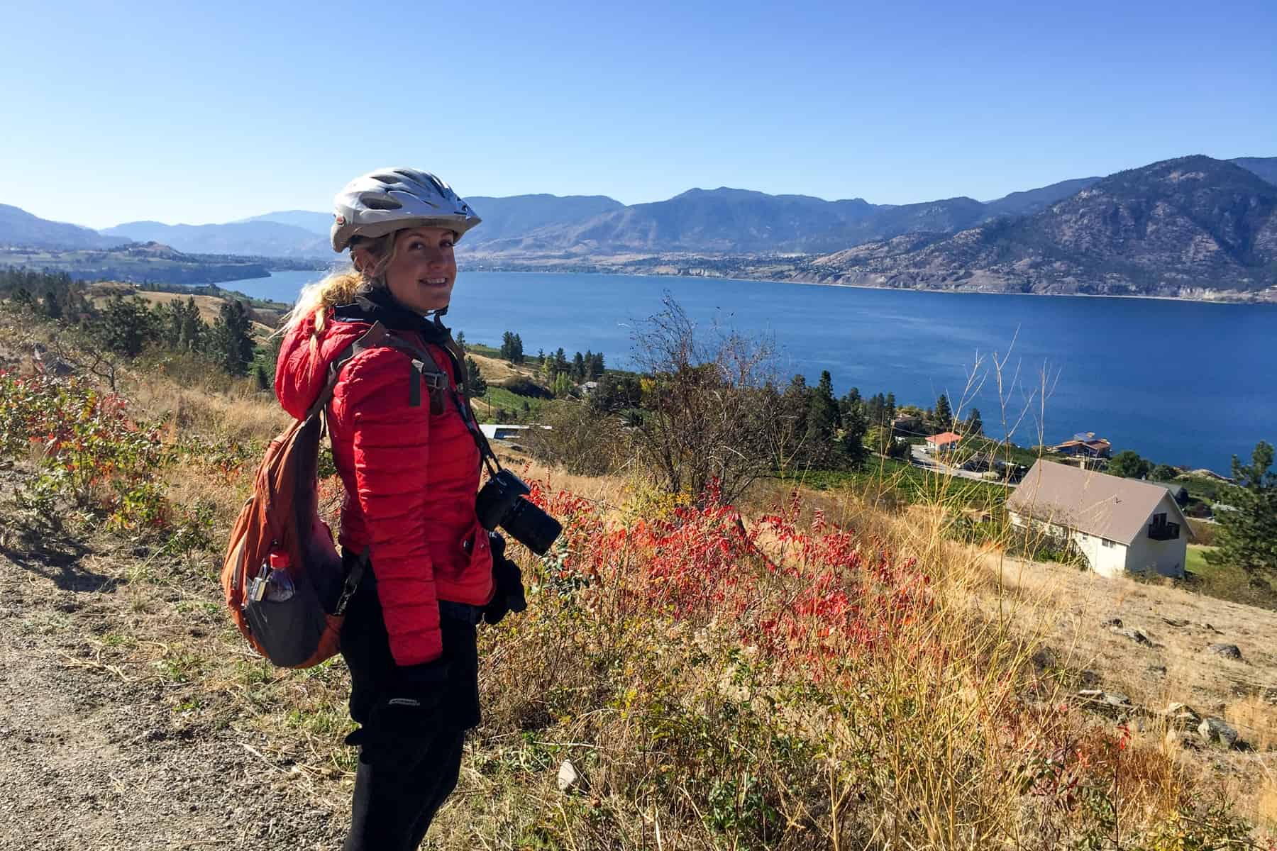 A woman in a bicycle helmet and with a camera around her neck, stands on the top of a small slope in the Kettle Valley overlooking the wide Kettle River. 