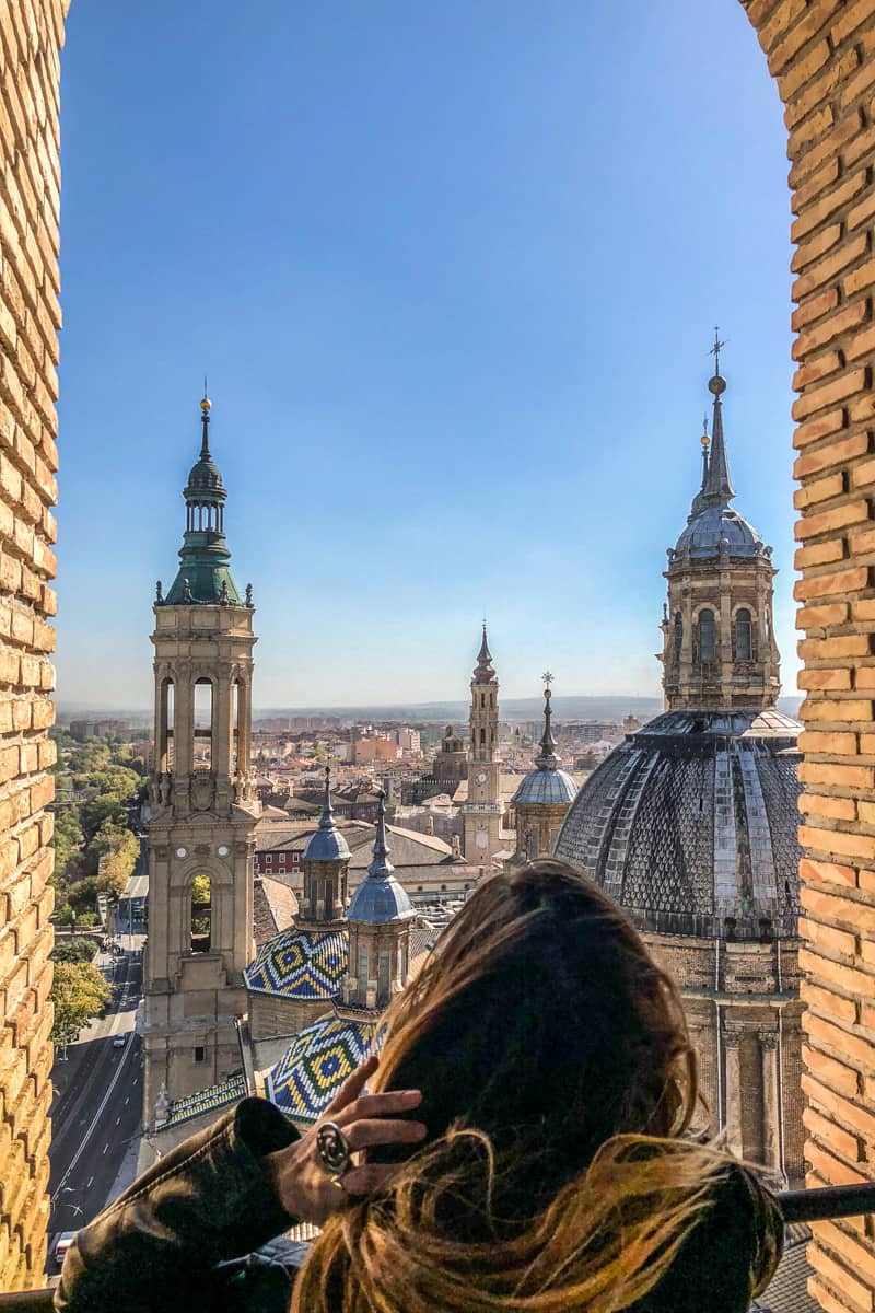 A woman holds her hair while looking out of a brick window archway towards a the city of Zaragoza's blue and yellow mosaic domed rooftops, spires and low-lying buildings. 