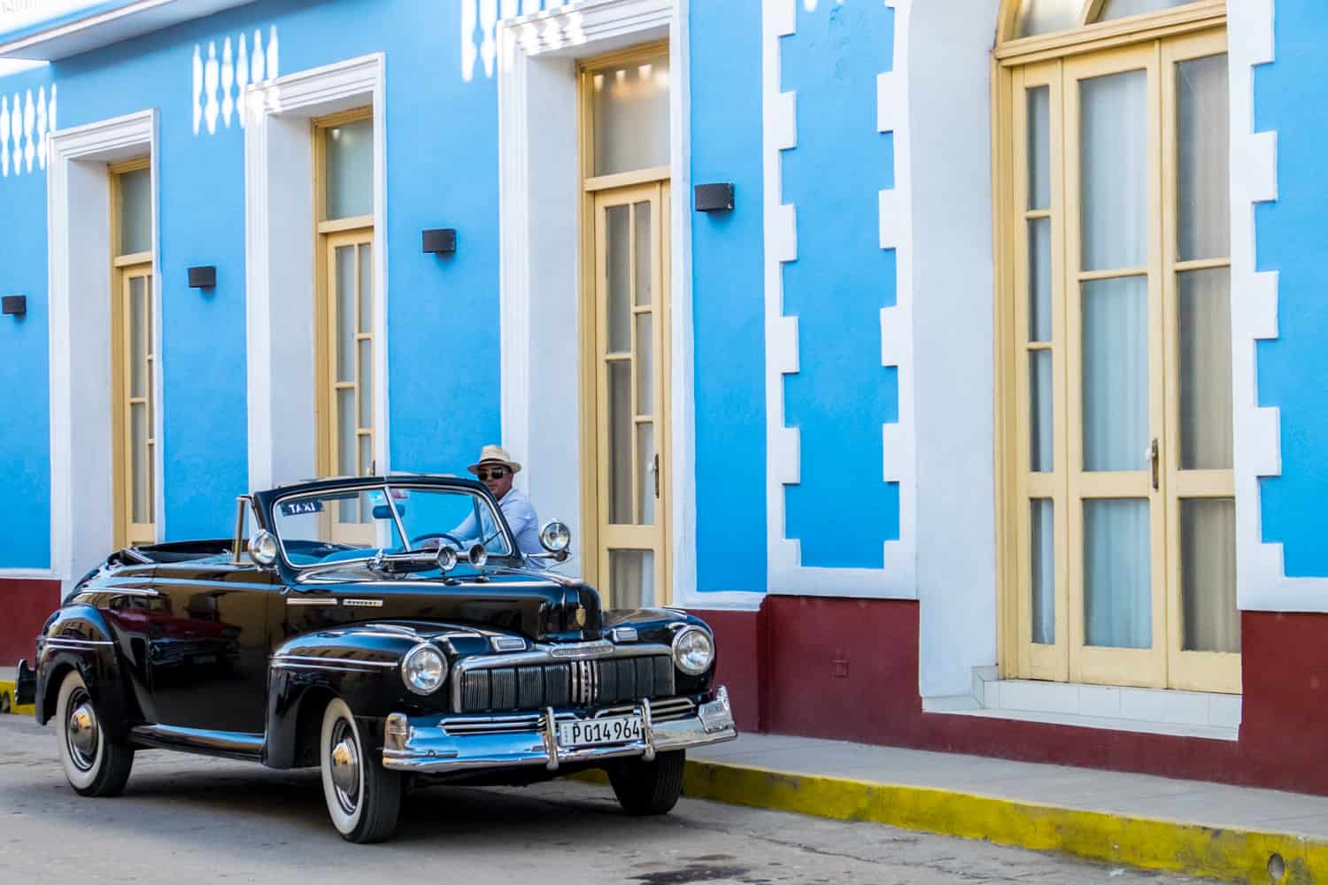 A man in a hat sits outside a blue and yellow house behind a black Cuban classic car taxi. 
