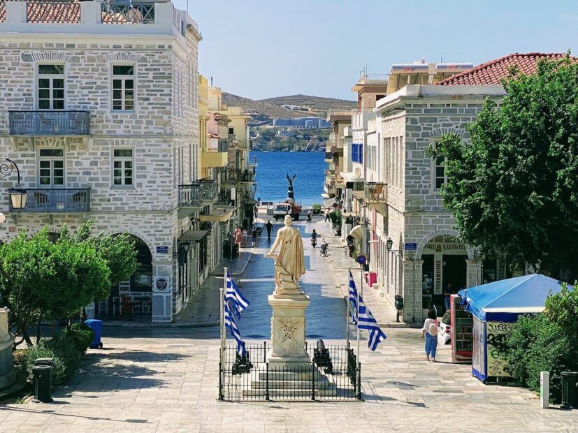 Main square with views of Greek flags, a statue and out to sea in Ermoupoli Syros Greece