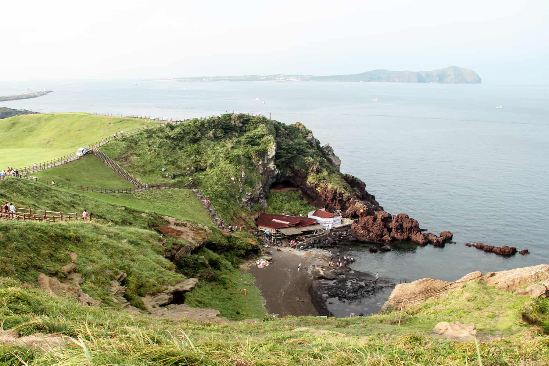 People strolling up a series of narrow walking paths on a green mound of Jeju Island's Sunrise Peak, looking down onto red-roofed houses, coastal rocks and the vast ocean