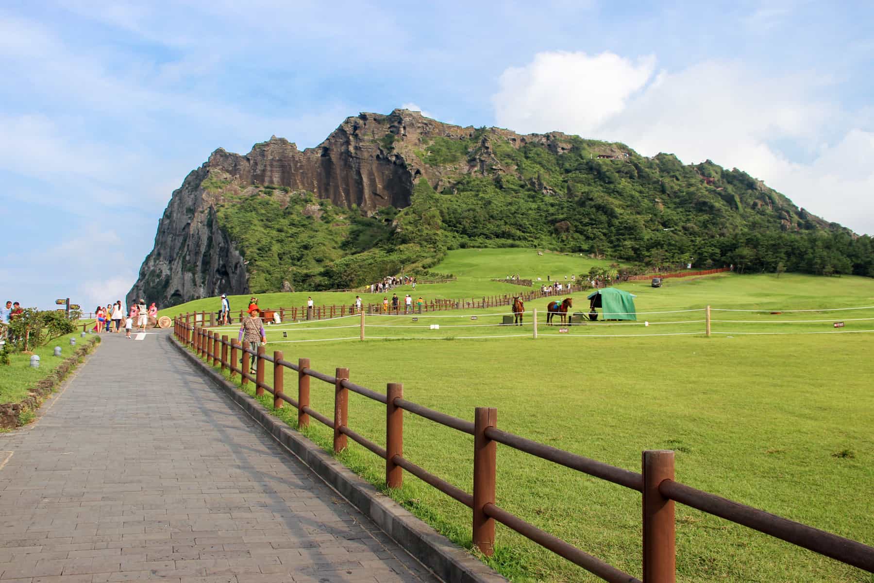 View looking towards a long path with red rails that leads to the green and brown volcanic mound of Seongsan ilchulbong in Jeju Island