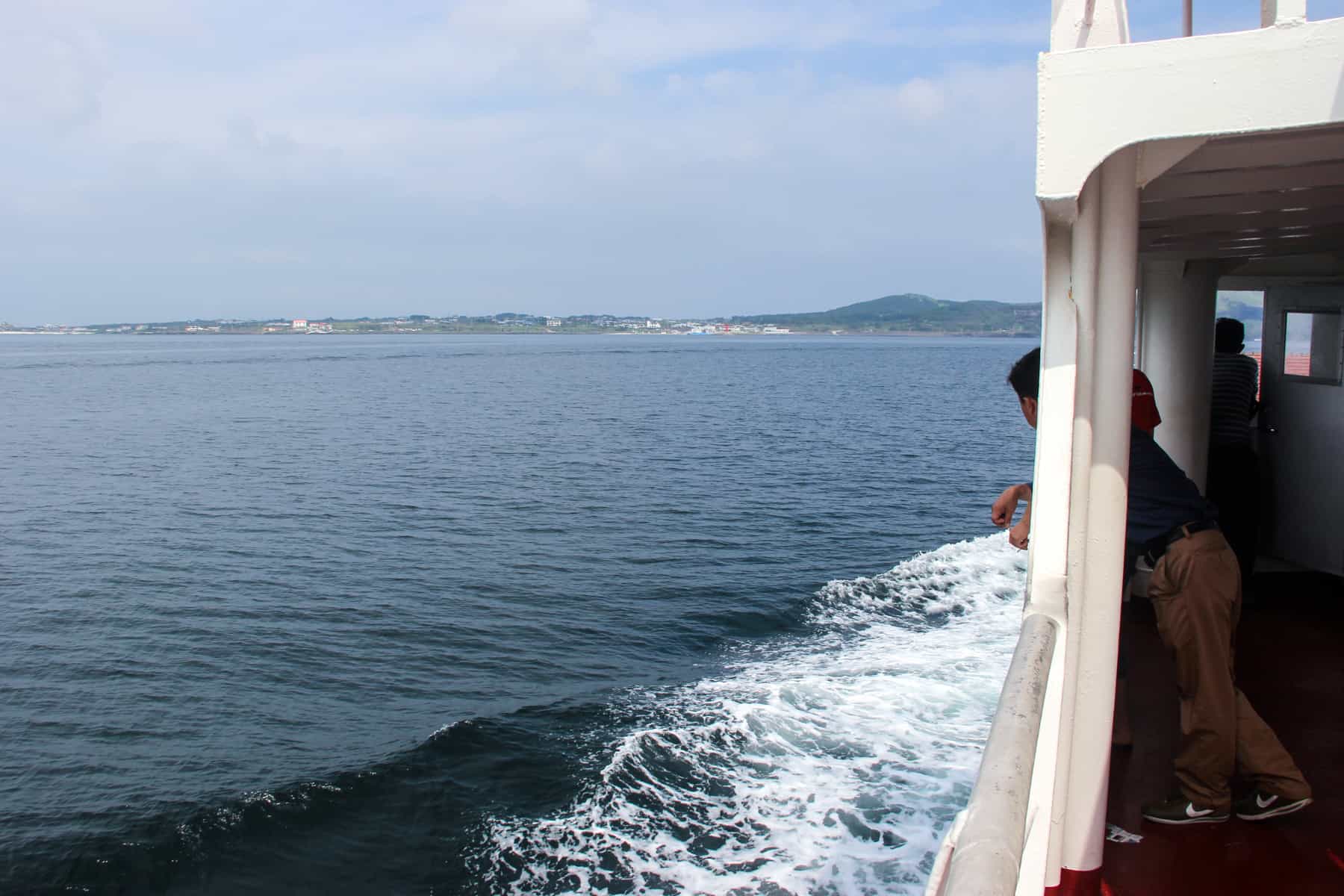 A man leans out from the side of a white ferry to Jeju Island Korea. The ocean is a grey blue and you can see the low volcanic peaks of the island in the background.