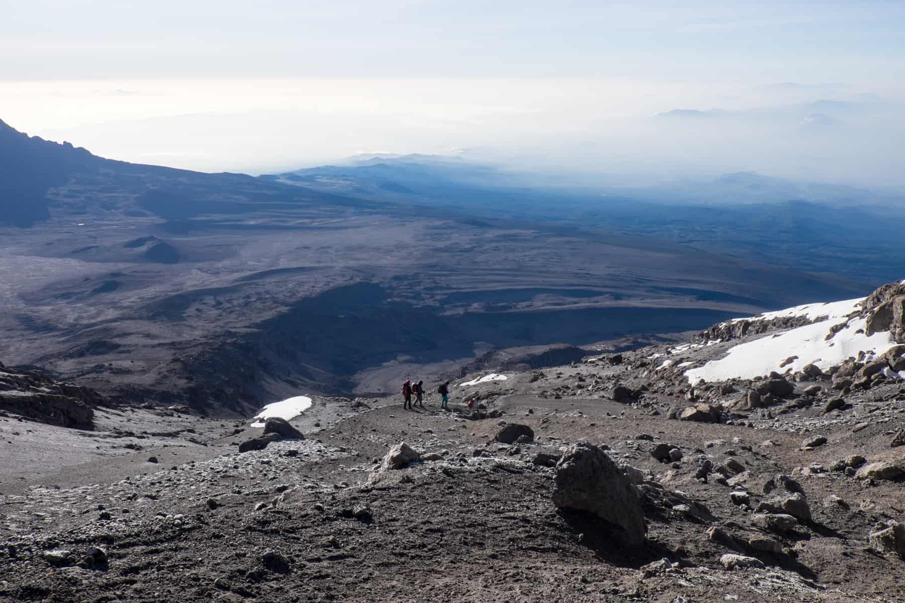 Three people, tiny in comparison to the wide rocky landscape, walk up a steep mountain slope towards a large patch of snow. In the background are more brown, rocky mountain slopes. 
