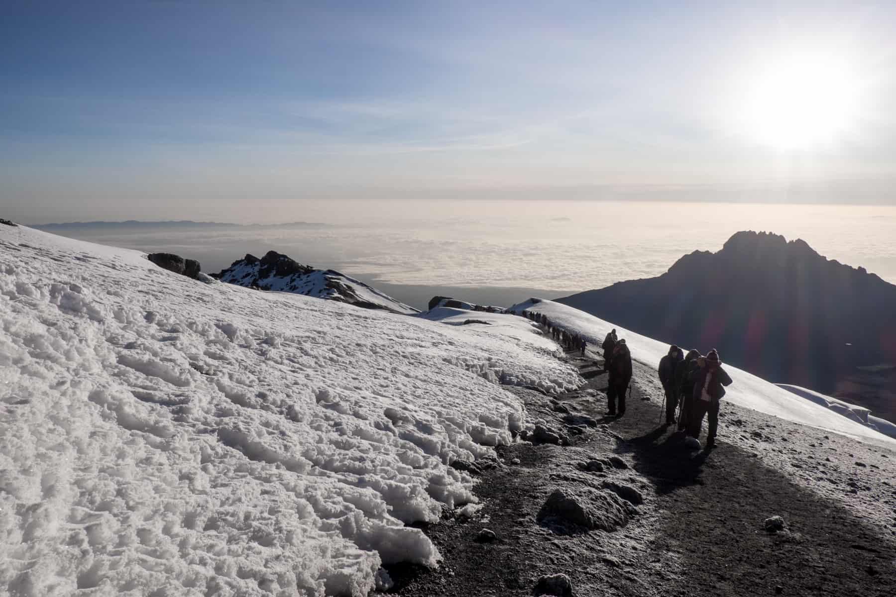 A line if people trek up the snow covered crater rim of Mt. Kilimanjaro. Behind them in a small peak and a seemingly never-ending layer of cloud. 