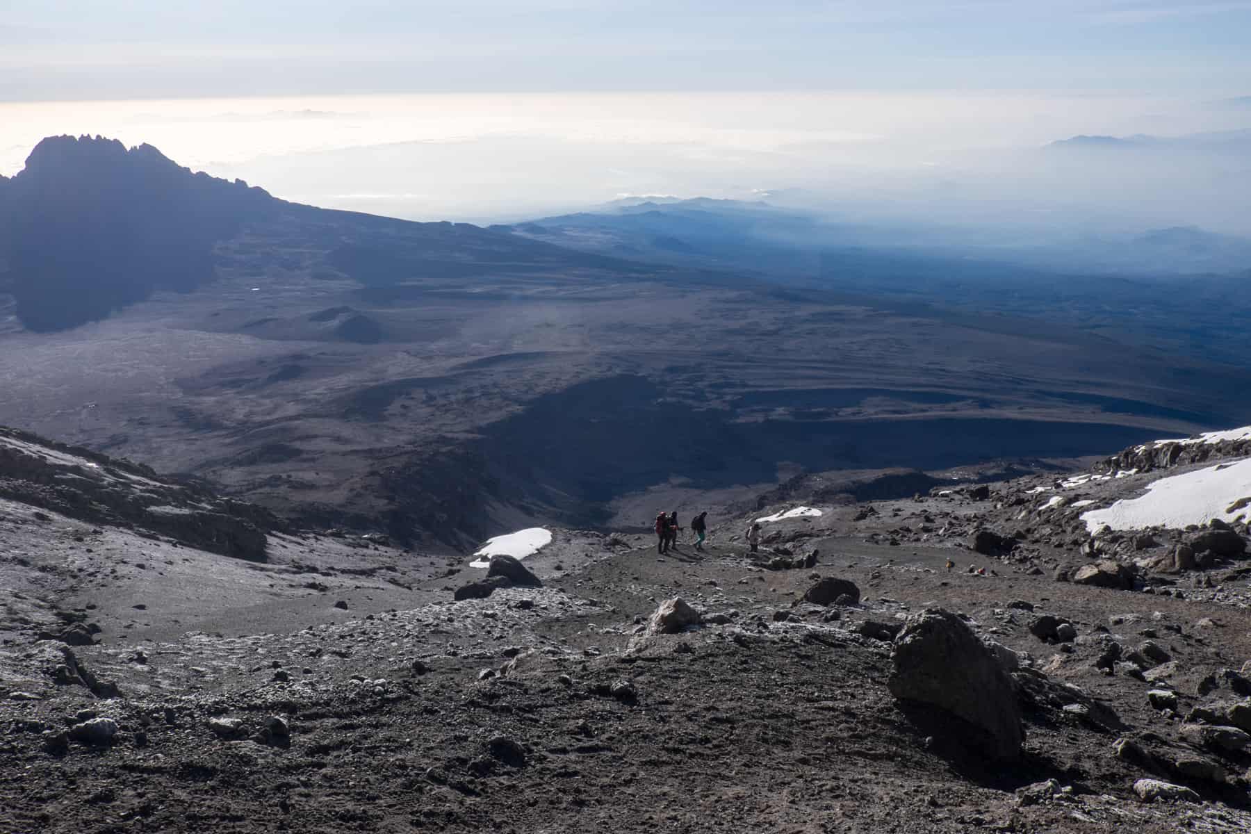 Four people climb up the dark, wide rocky pathway of Kilimanjaro's peak, backed by large volcanic slopes and small patches of melting snow.