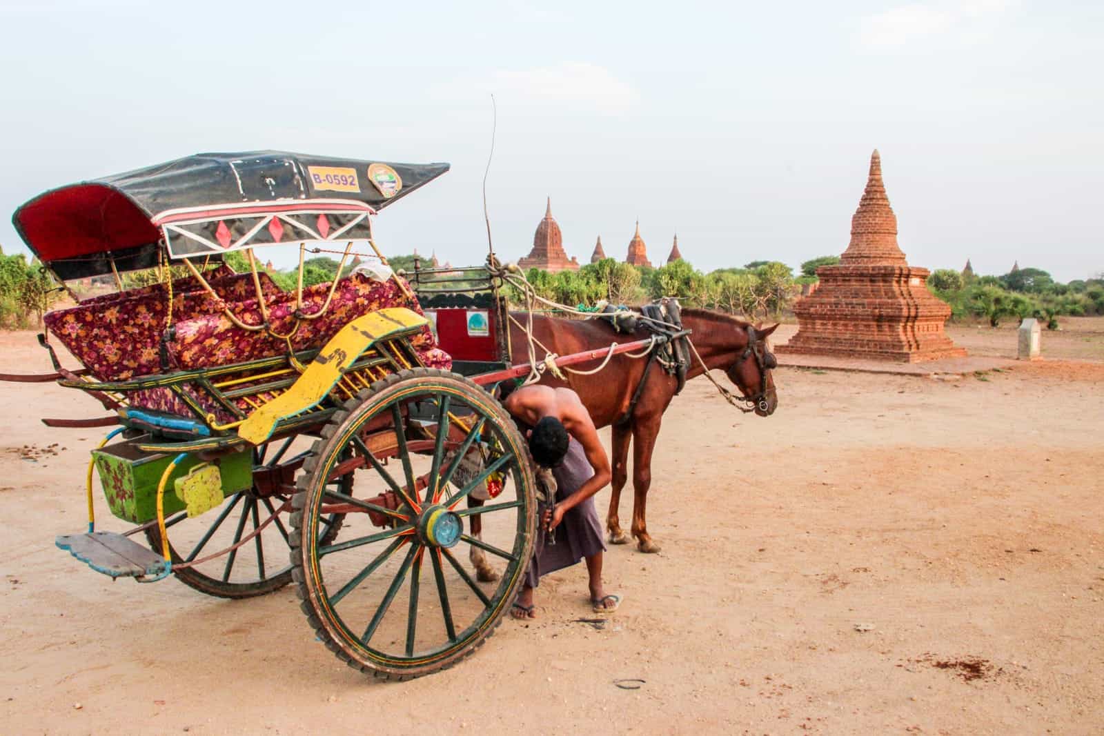 A topless, male horse and cart driver in a purple tunic skirt, bends down to fix the wheel of his red and yellow cart in Bagan, Myanmar. Five golden brown temples can be seen behind him. 