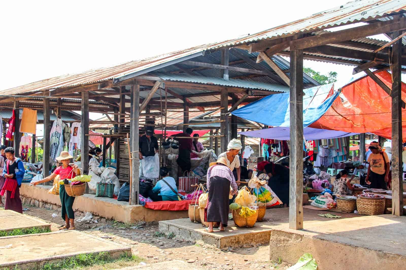 Two women tie bags of shopping onto a wooden carrying pole. They stand within an open spot of a busy covered market place in Myanmar full of people and goods.