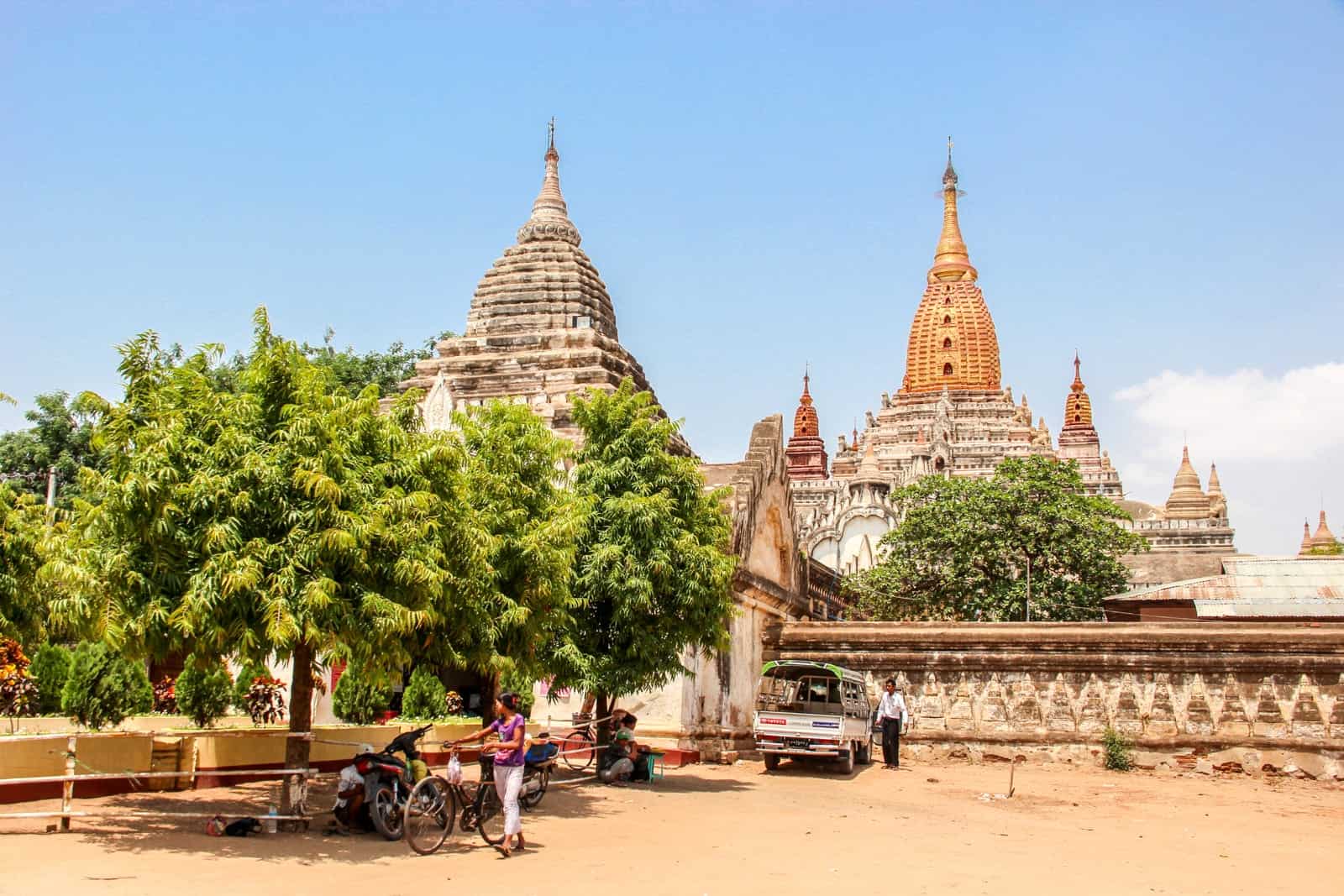 A woman in a pink t-shirt and white trousers pushes a bike in front of a tree at the entrance to a large temple complex with white and gold pagodas, in the dusty orange grounds of Bagan, Myanmar