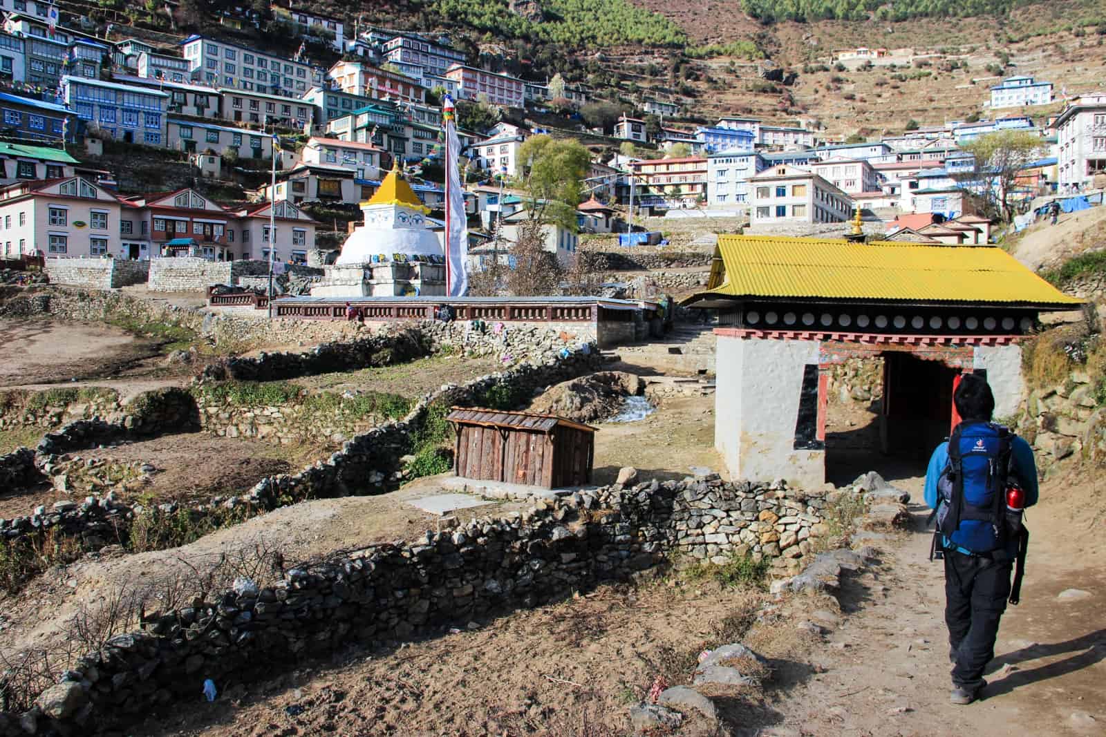 A man with a blue backpack walked towards a white gate with a yellow roof - the entrance to the Namche Bazaar village on the EBC Trek - towards a small white and gold temple structure and a cluster of and a cluster of rectangular houses built on the hillside
