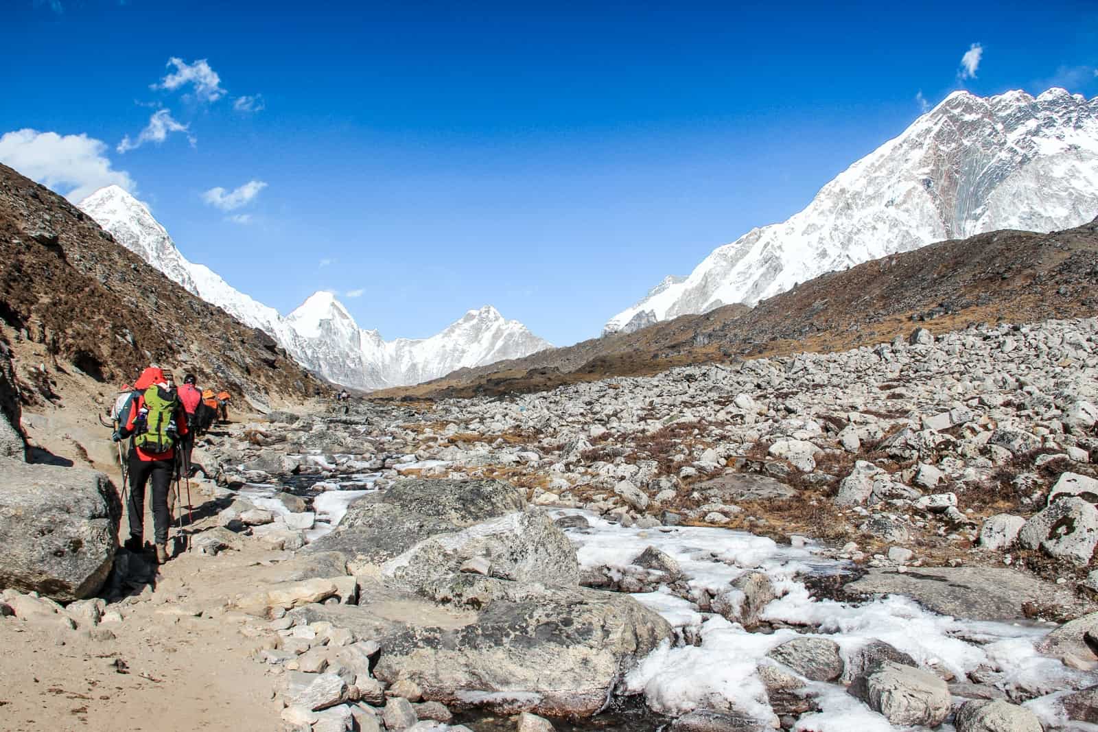A small line of trekker walk on a nasty, dusty path past a wide rocky scree on the Everest Base Camp Trek. The dry, brown valley is surrounded by bright, white mountain ranges. 