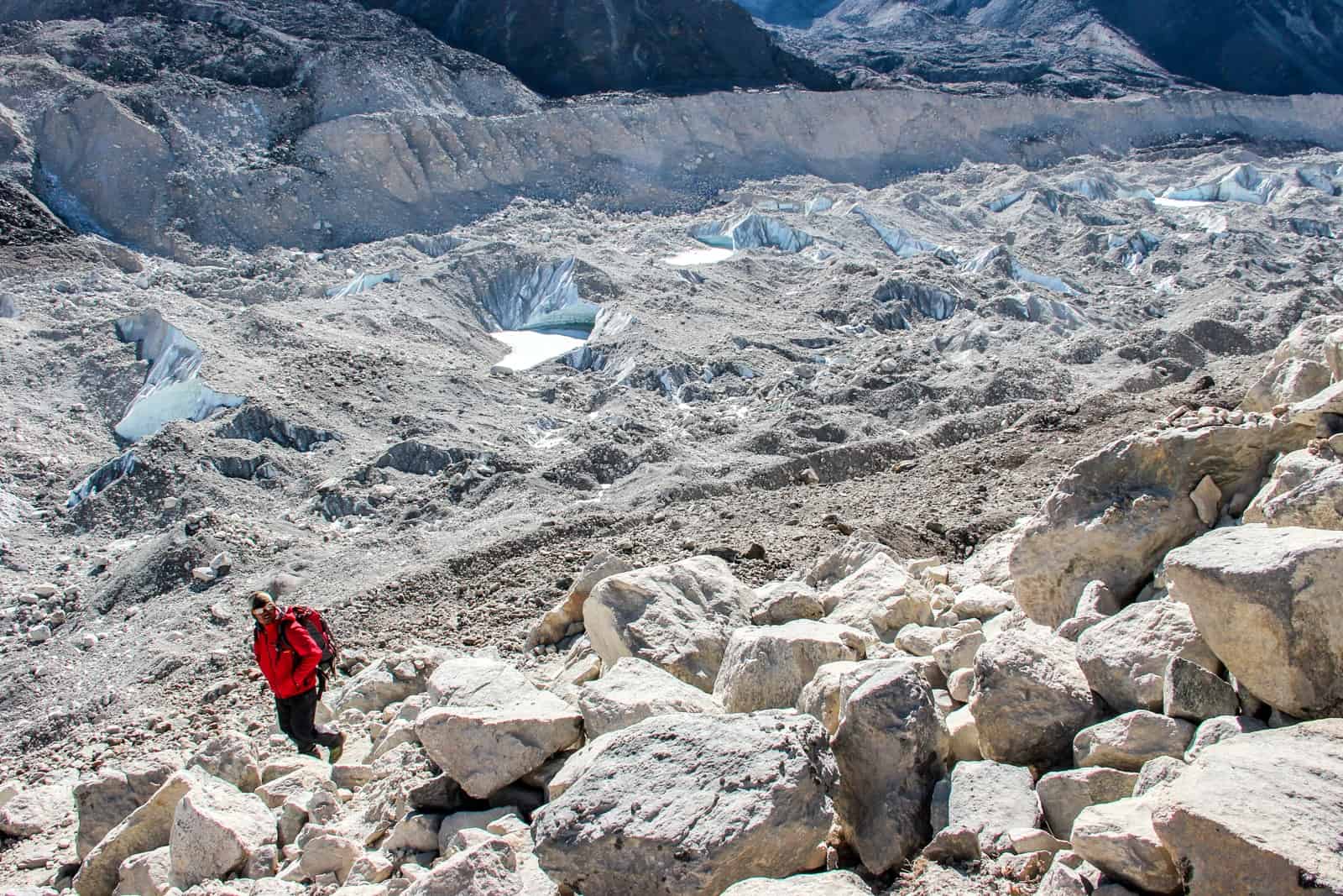 A Everest Base camp trek guide in a red jacket walks past huge sand coloured boulders on one side, and pockets of ice in silver dust on the other. 