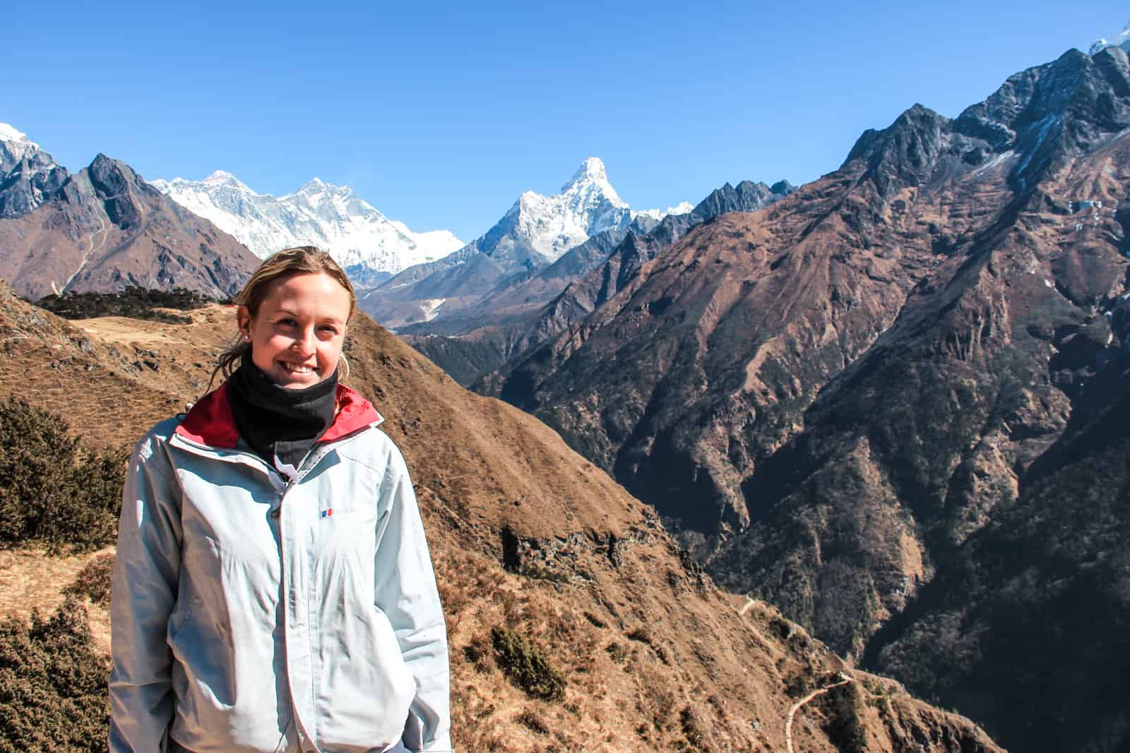 A smiling woman in a pale blue jacket stands on a golden brown mountain hillside. In the far distance is the snow capped Mount Everest