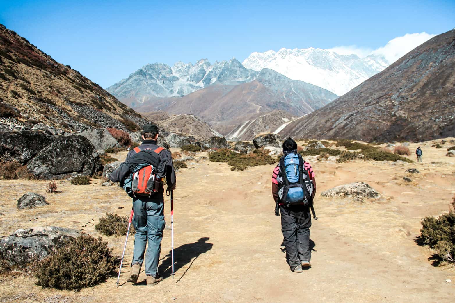 Two men with backpacks walk through the yellow dusty pathway of a wide mountain valley on the Everest Base Camp Trek. They walk towards a grey and white snow-capped mountain ridge, between two brown peaks. 