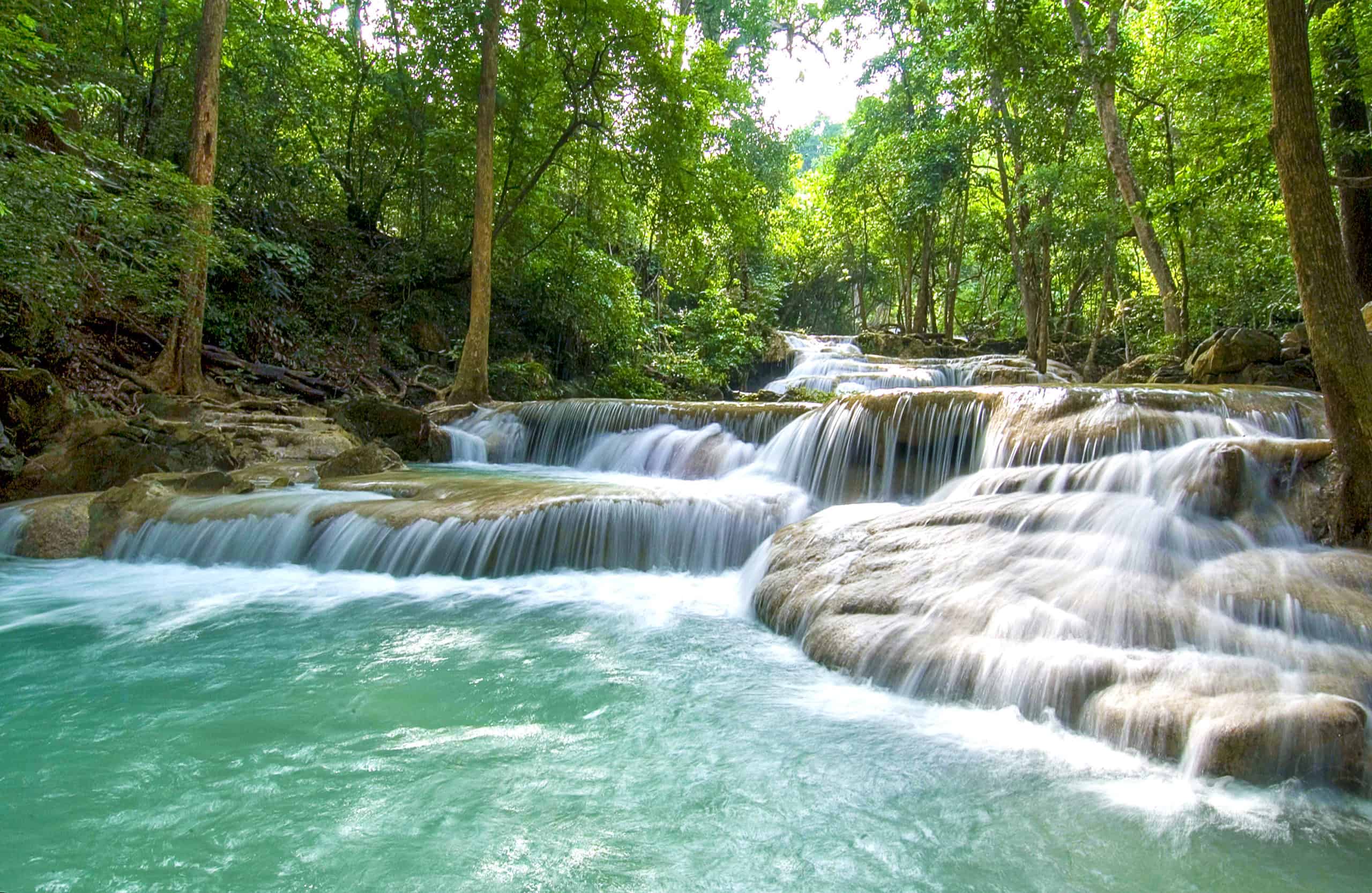 Tiered waterfalls with turquoise water pools within a dense forest - part of the Erawan Waterfalls in Kanchanaburi, Thailand.