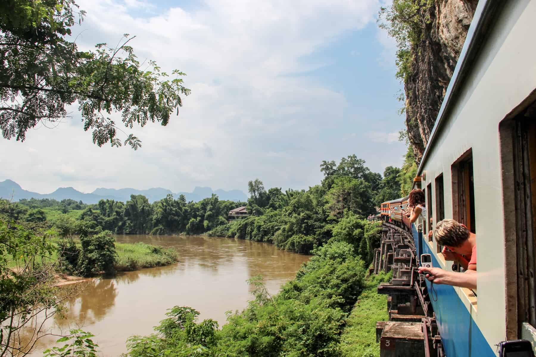People lean out of a blue and white train - the Death Railway in Kanchanaburi – as it curves past a jungle green river bank
