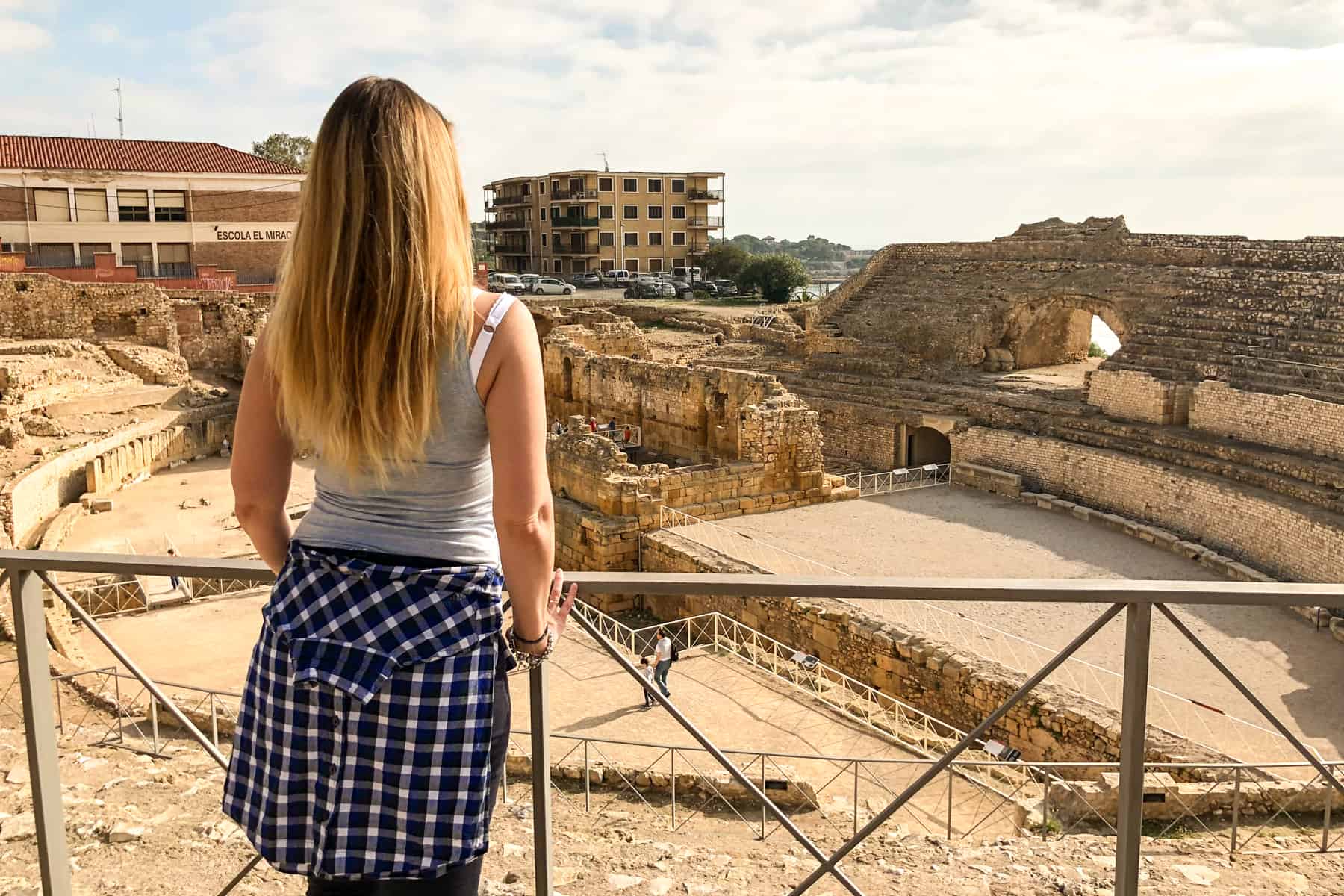A woman with long blonde hair stands at a metal rail overlooking the romans ruins of the Tarragona Amphitheatre in Spain 