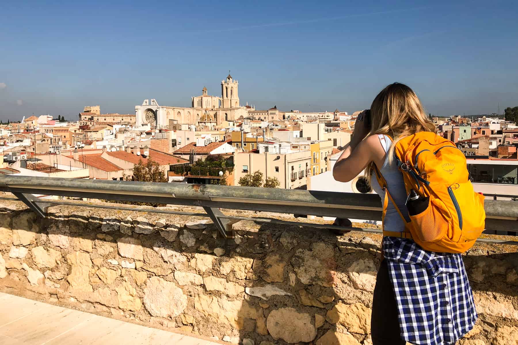 A woman wearing a yellow backpack takes a picture of the city of Tarragona from the top of the golden stone Roman Circus building