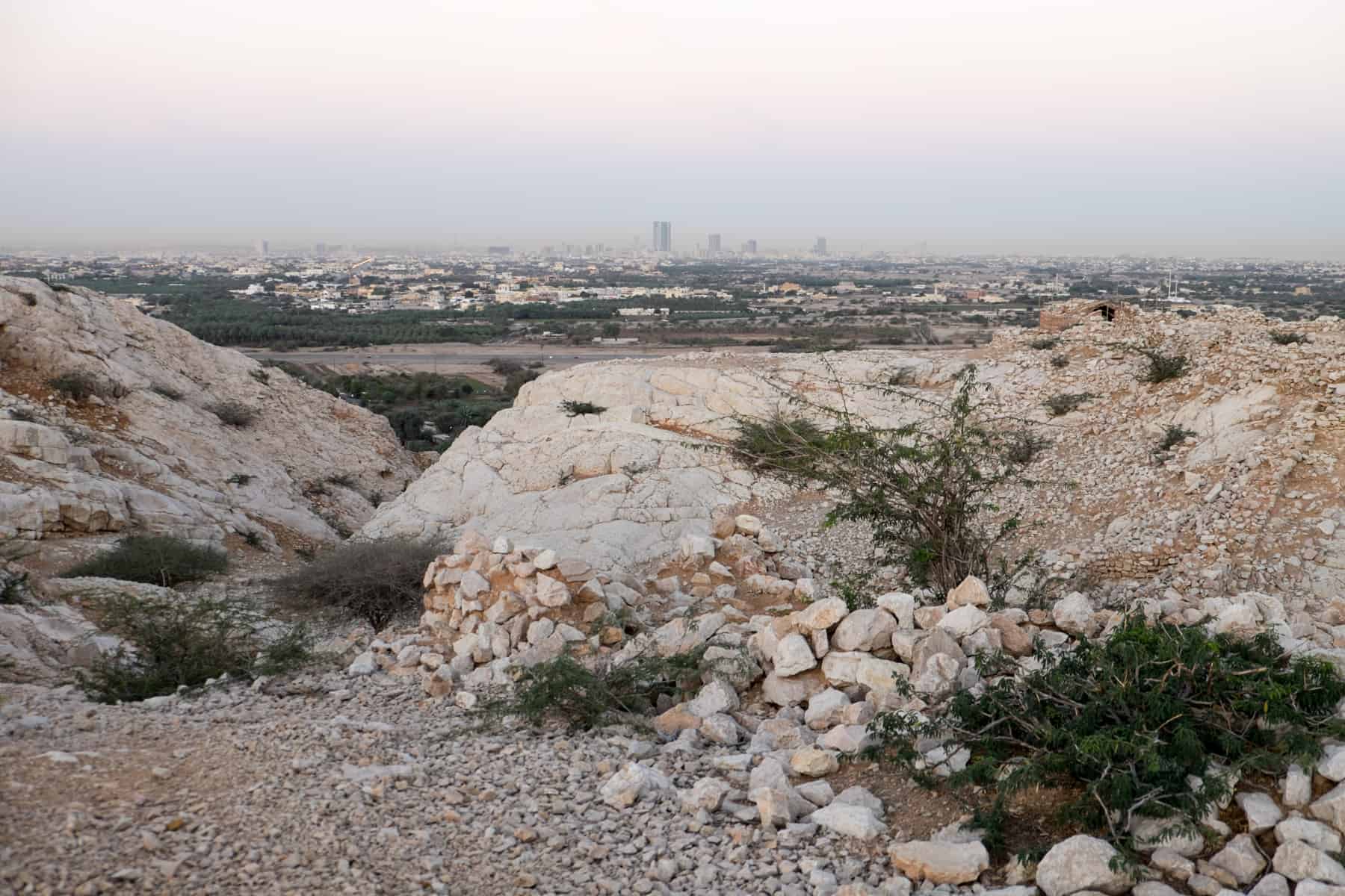 Mounds of smooth white stones and pebbles on a hilltop - the ruins of Sheba's Palace - overlook the modern skyline of Ras Al Khaimah city in the background