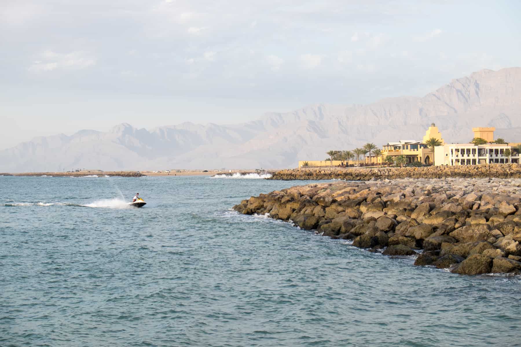 A man on a jetski on grey-blue waters ride close to a rocky wall on a coastline backed by a golden yellow resort and brown mountains