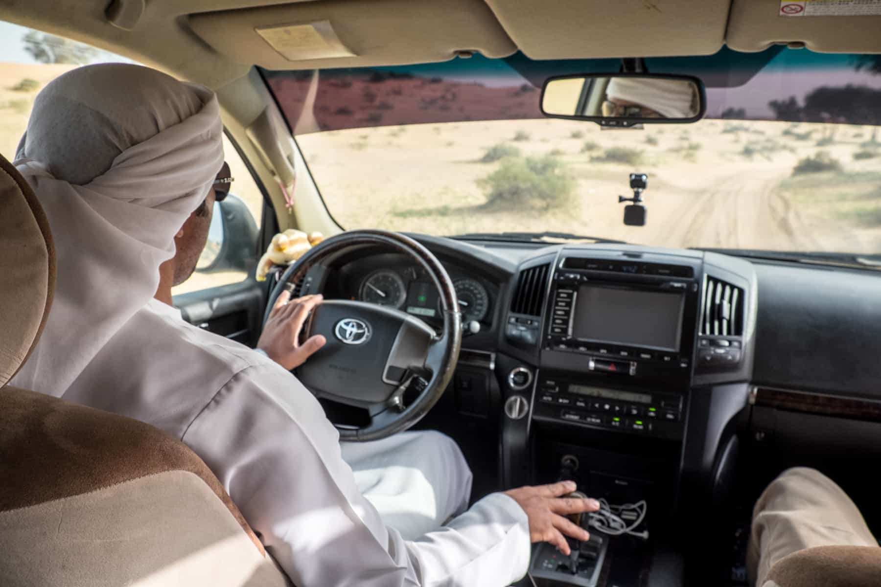 A man wearing a white robe and head scarf drives a beige and black car through a desert landscape