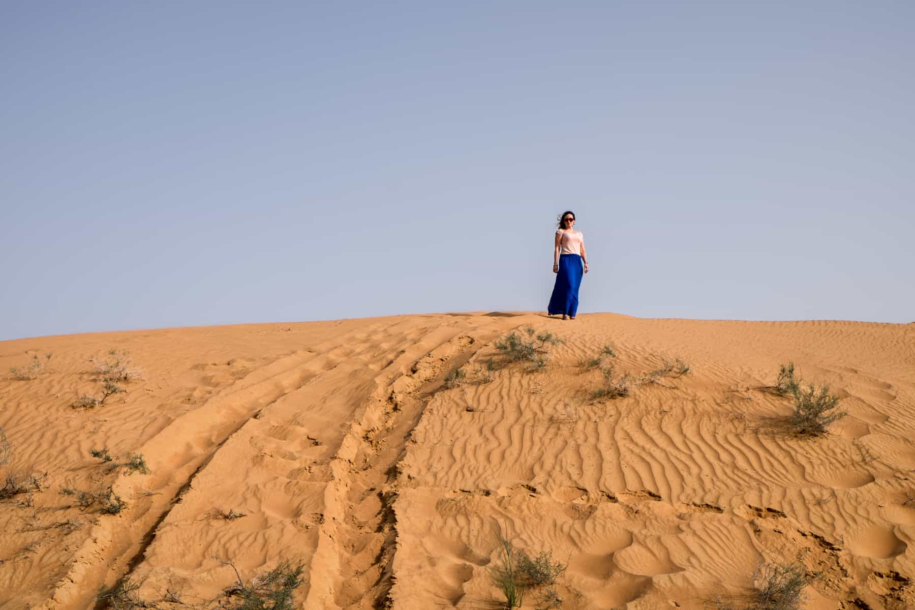 A woman in a pink t-shirt and electric blue skirt stands on top of a golden sand dune in Ras Al Khaimah, under a clear blue sky